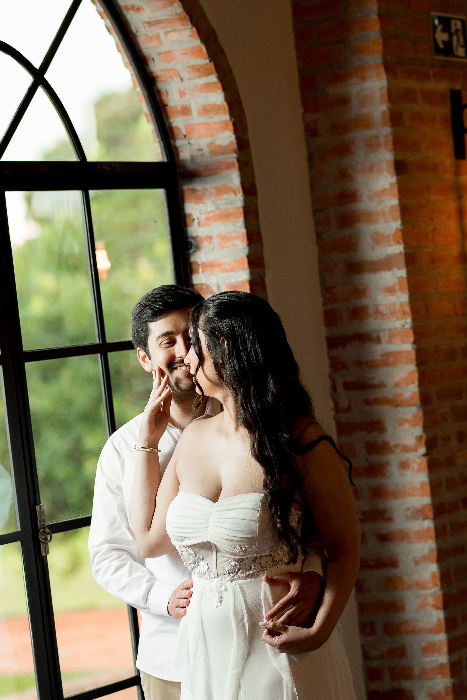 Bride and groom share a romantic moment by an arched window with brick walls in a wedding venue.