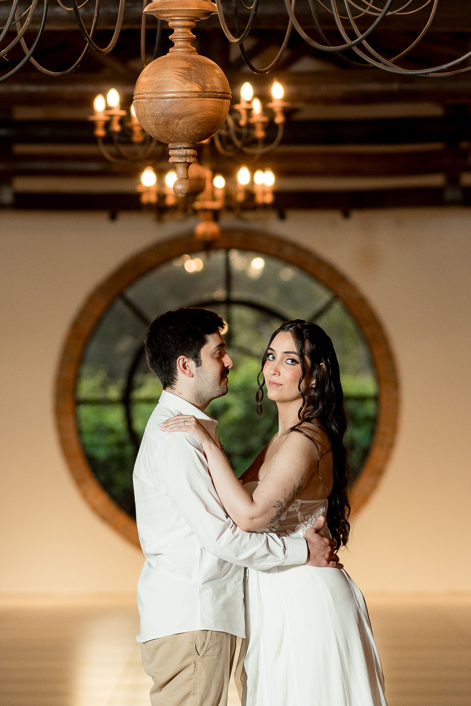 Couple embracing in elegant venue with round window and chandelier, bride in white gown gazing at camera.