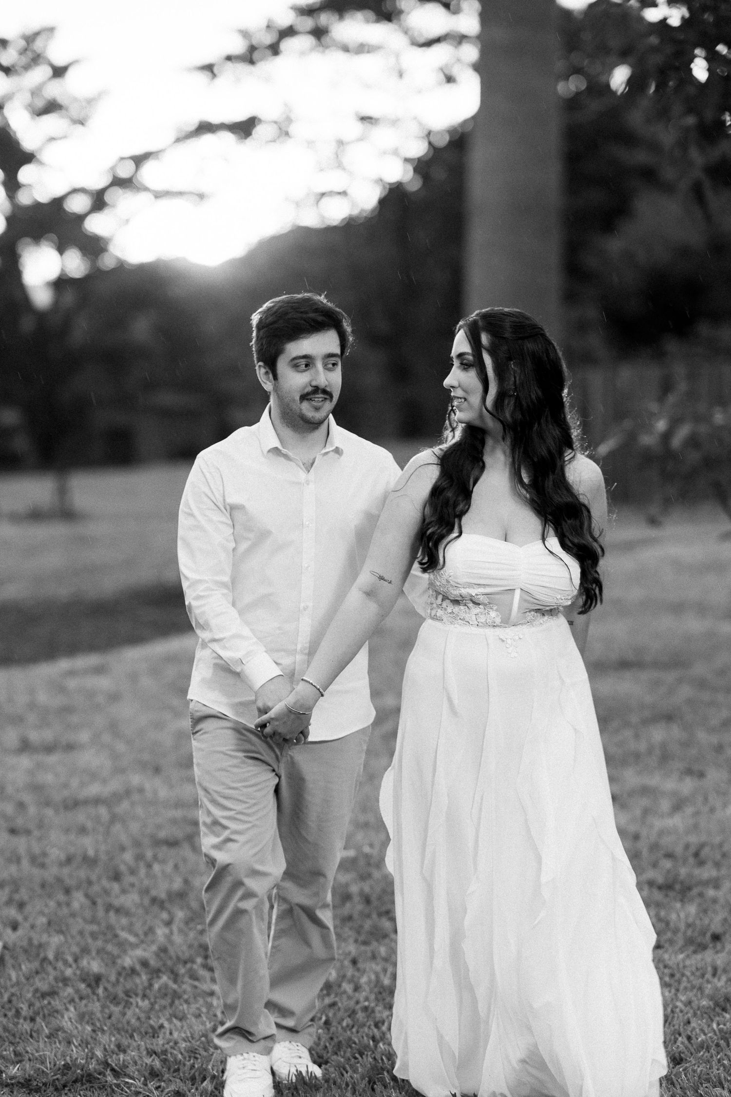 Black and white photo of a couple holding hands in a park, woman in white gown, man in casual attire at sunset.