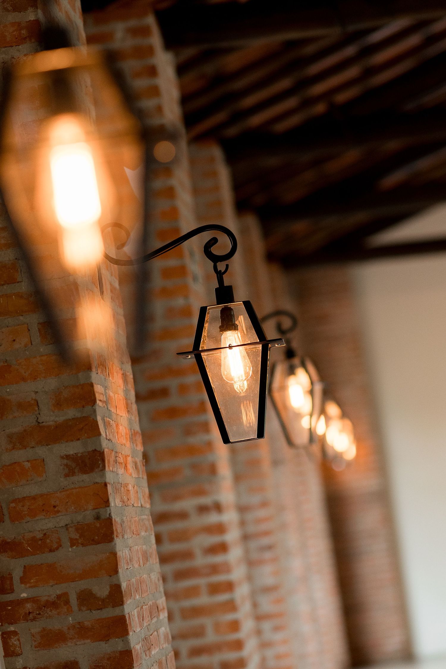 Row of vintage black metal lanterns with Edison bulbs hanging on rustic brick wall under wooden ceiling.