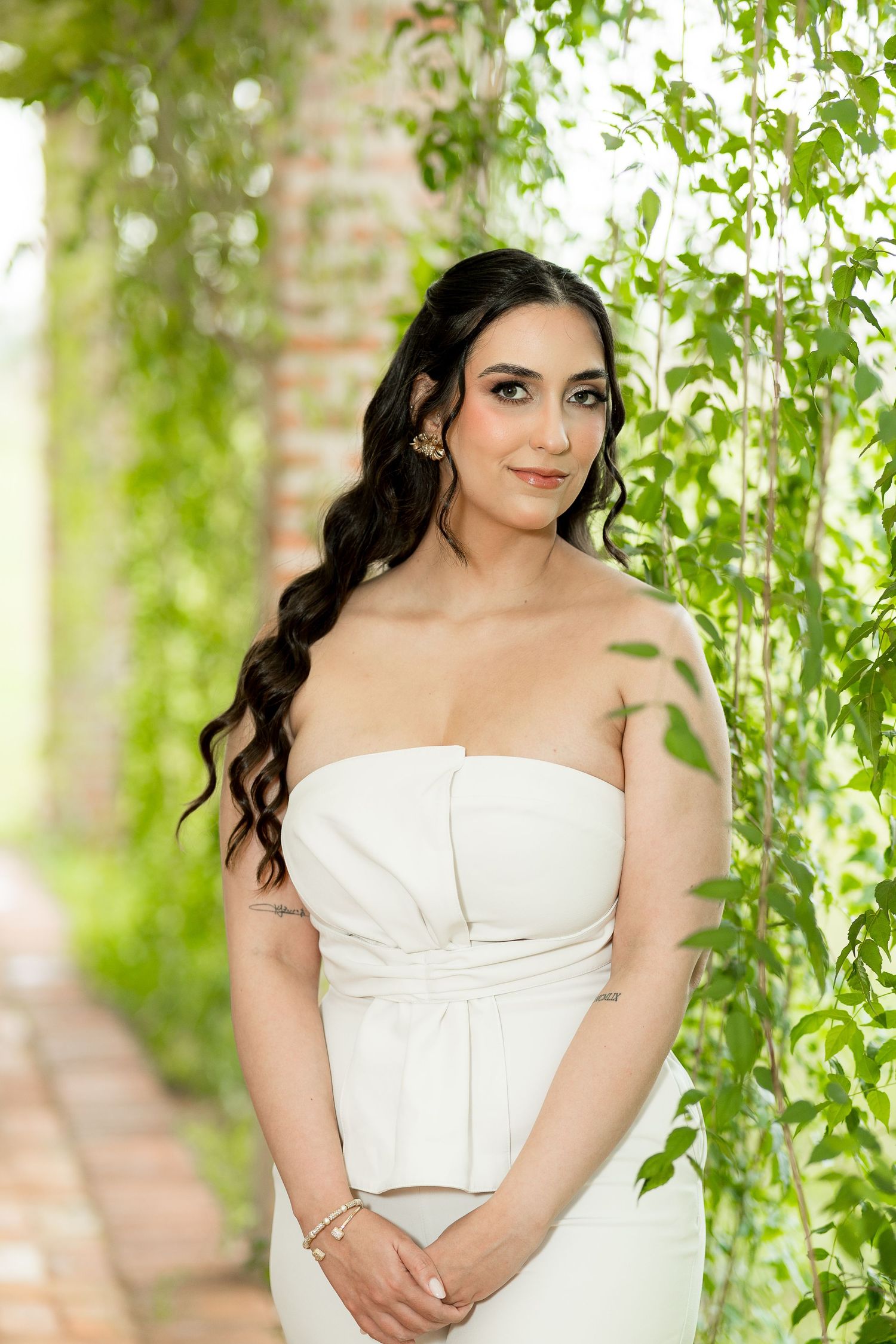 Bride in white strapless gown poses gracefully among lush green vines and brick columns.