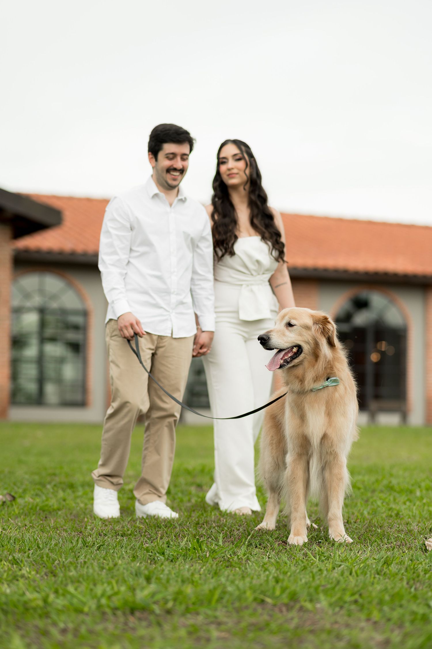 Smiling couple in white outfits walking a golden retriever on a leash on green grass near a building.