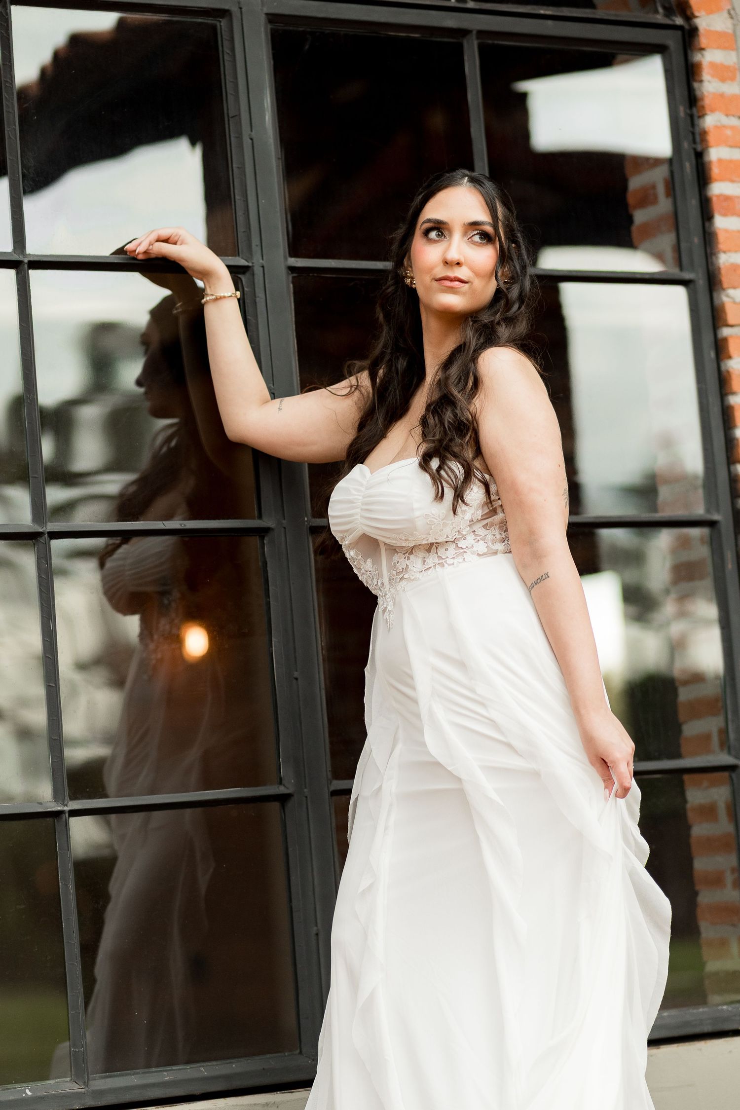 Bride in white strapless gown poses gracefully by black-framed windows, her reflection visible in the glass.