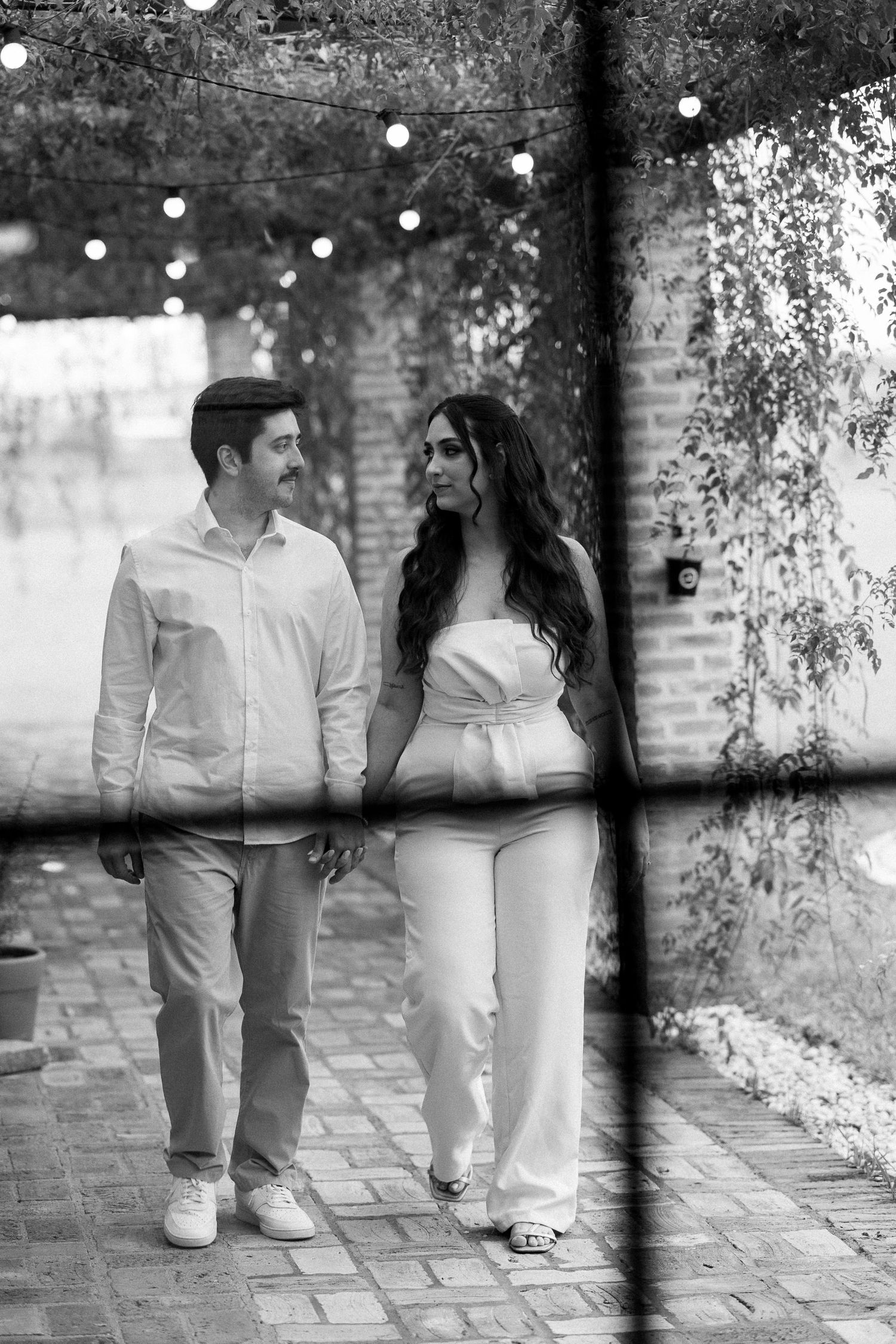 Black and white photo of a couple walking hand in hand on a brick path with string lights and trees in the background.