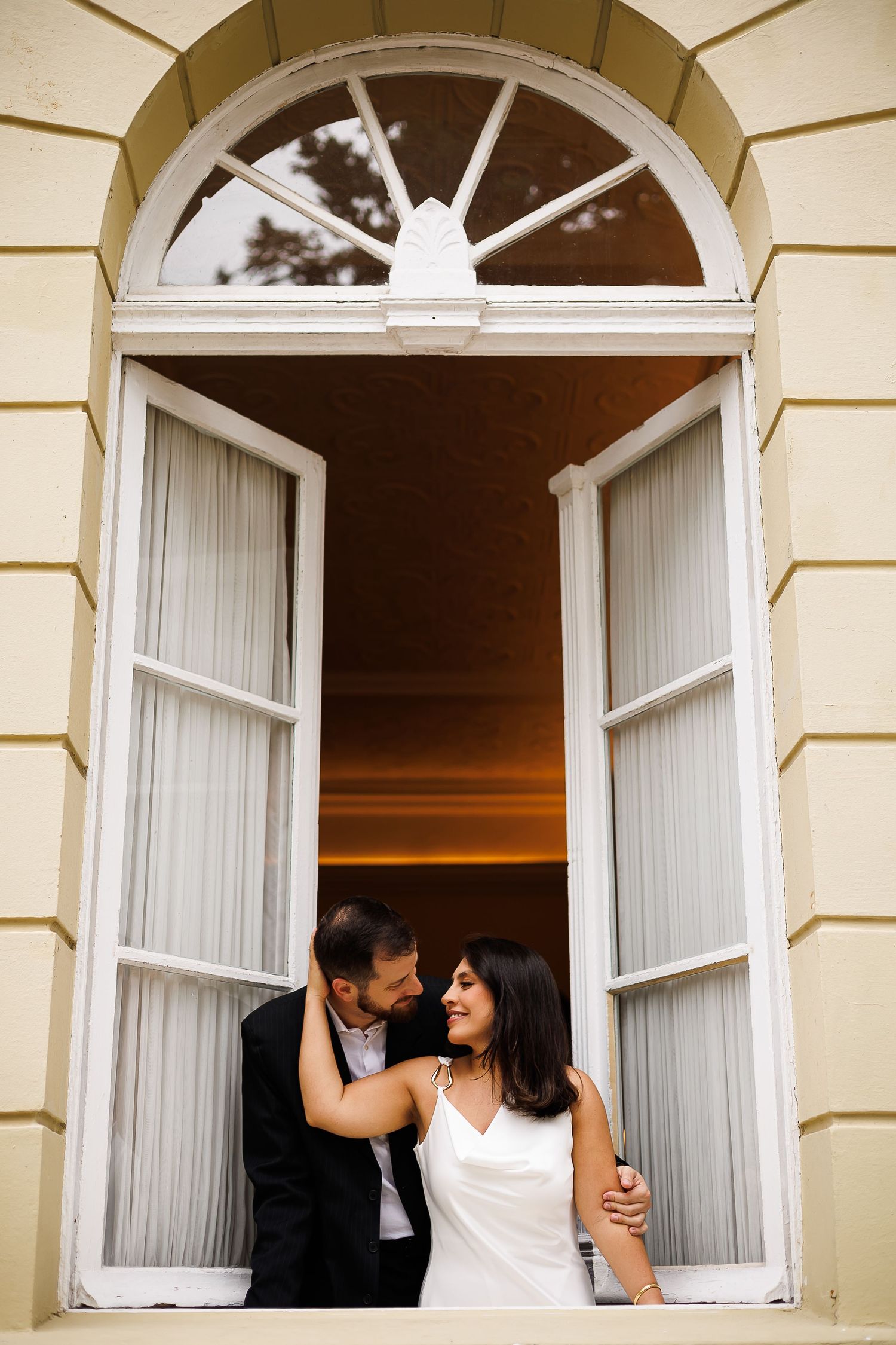 Couple in wedding attire sharing a romantic moment in an elegant arched window at sunset.