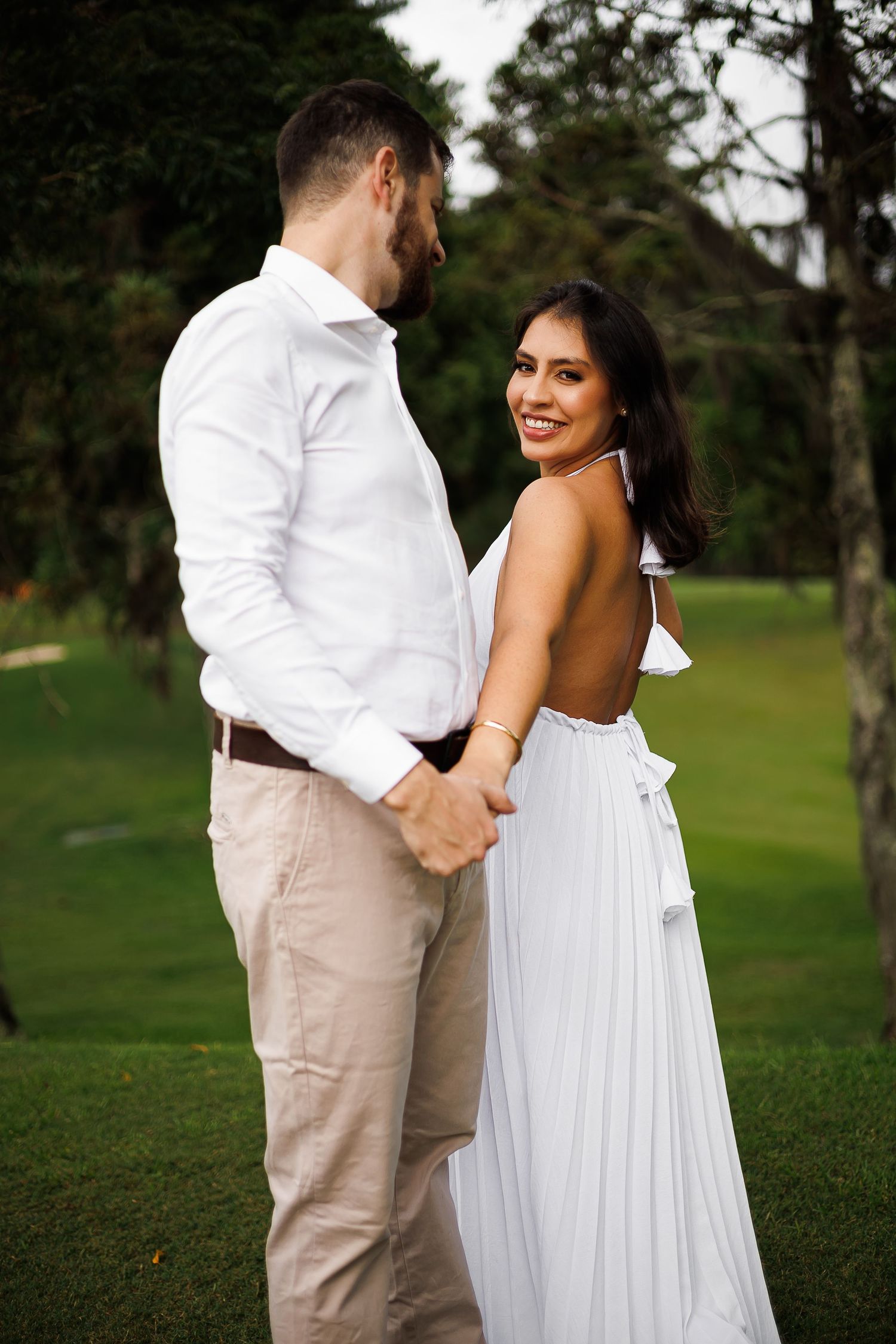 A smiling woman in a white backless dress holds hands with a man in a white shirt outdoors.