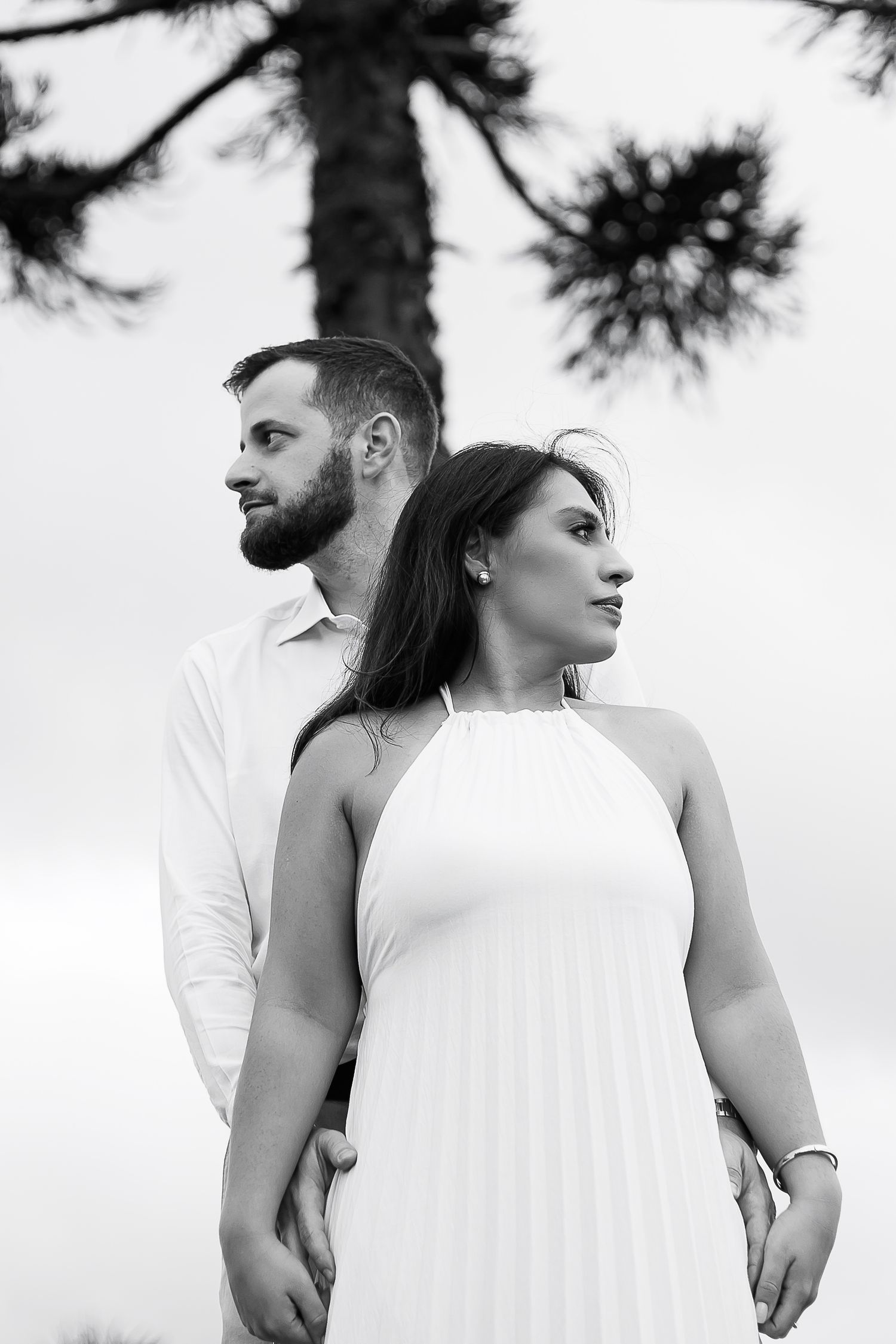 Black and white portrait of a couple standing back to back outdoors near a pine tree, woman in white dress.
