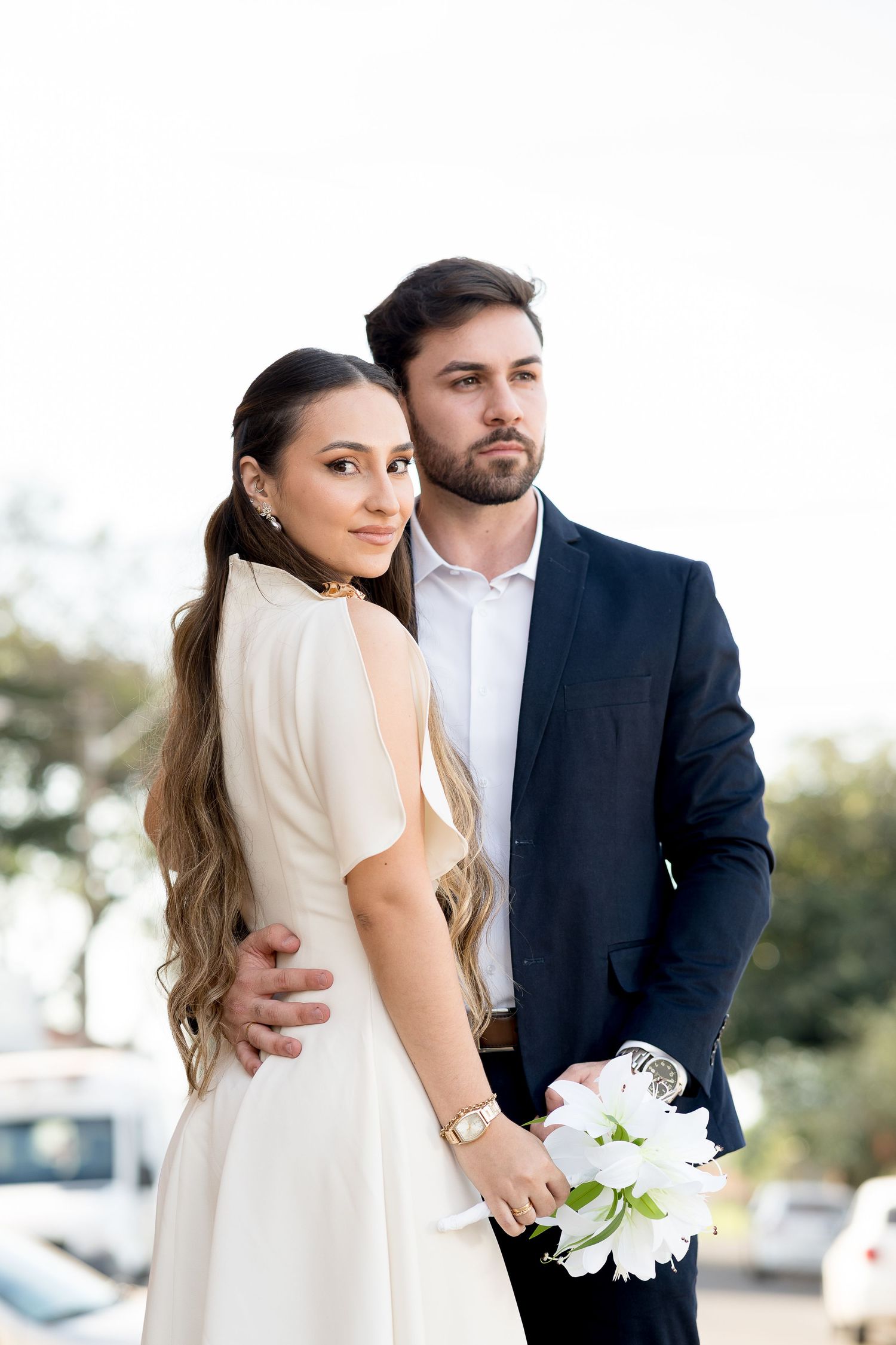 Elegant couple in wedding attire, bride in white dress holding white lilies, groom in navy suit embracing outdoors.