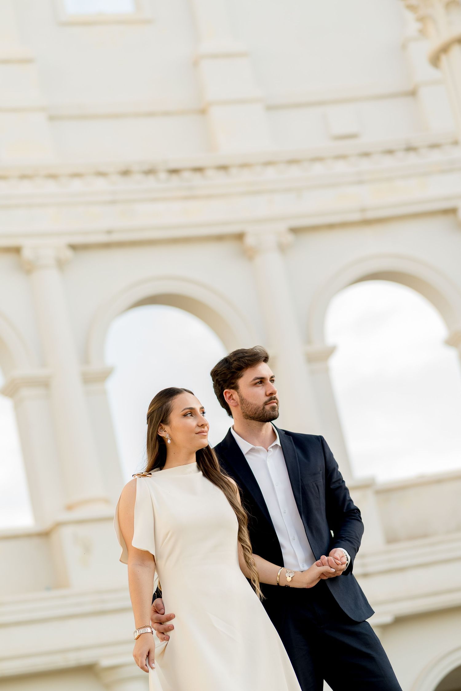 Elegant couple holding hands in front of the Roman Colosseum, woman in white dress, man in navy suit.