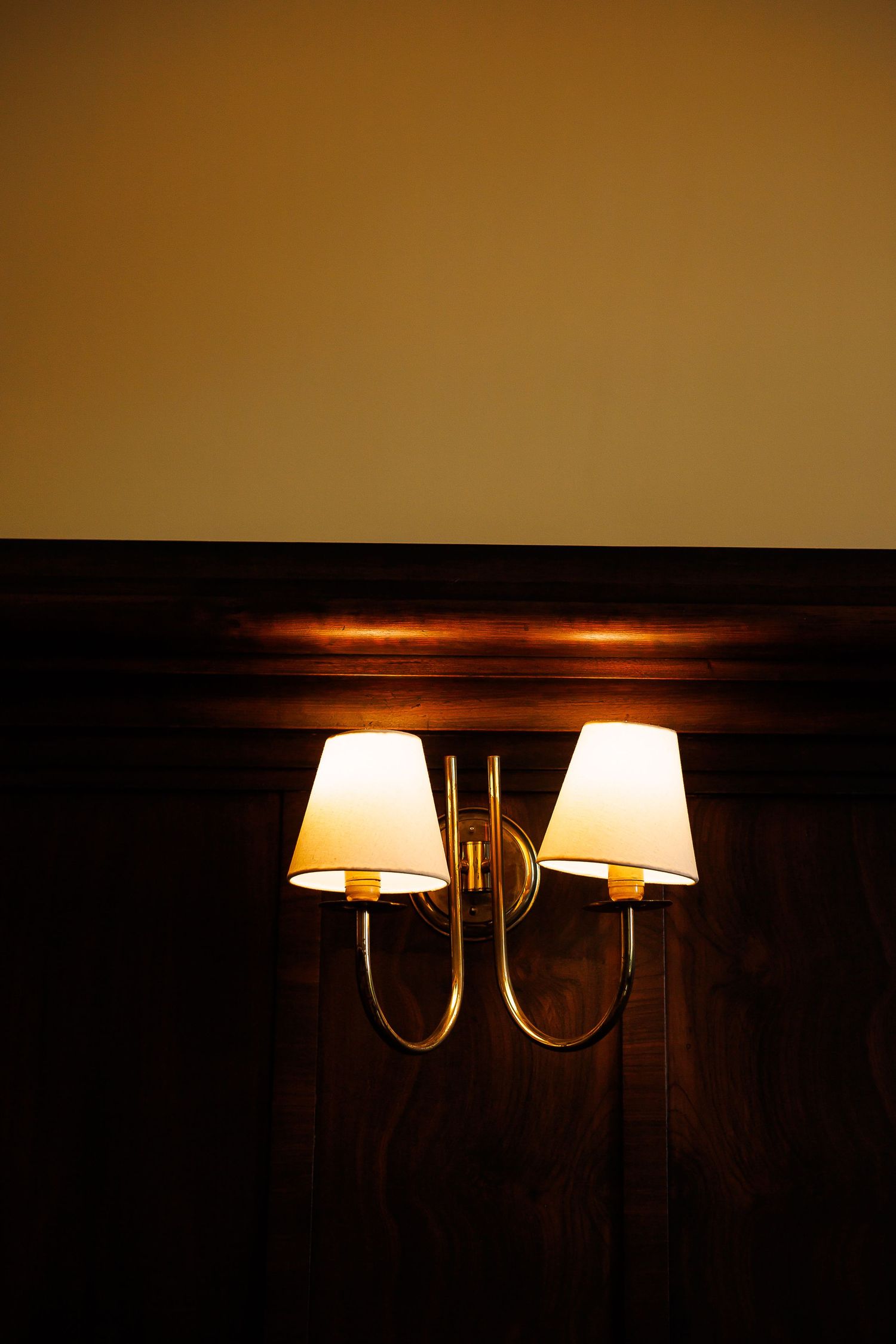 Elegant double wall sconce with brass finish and white shades glowing warmly against dark wood paneling.
