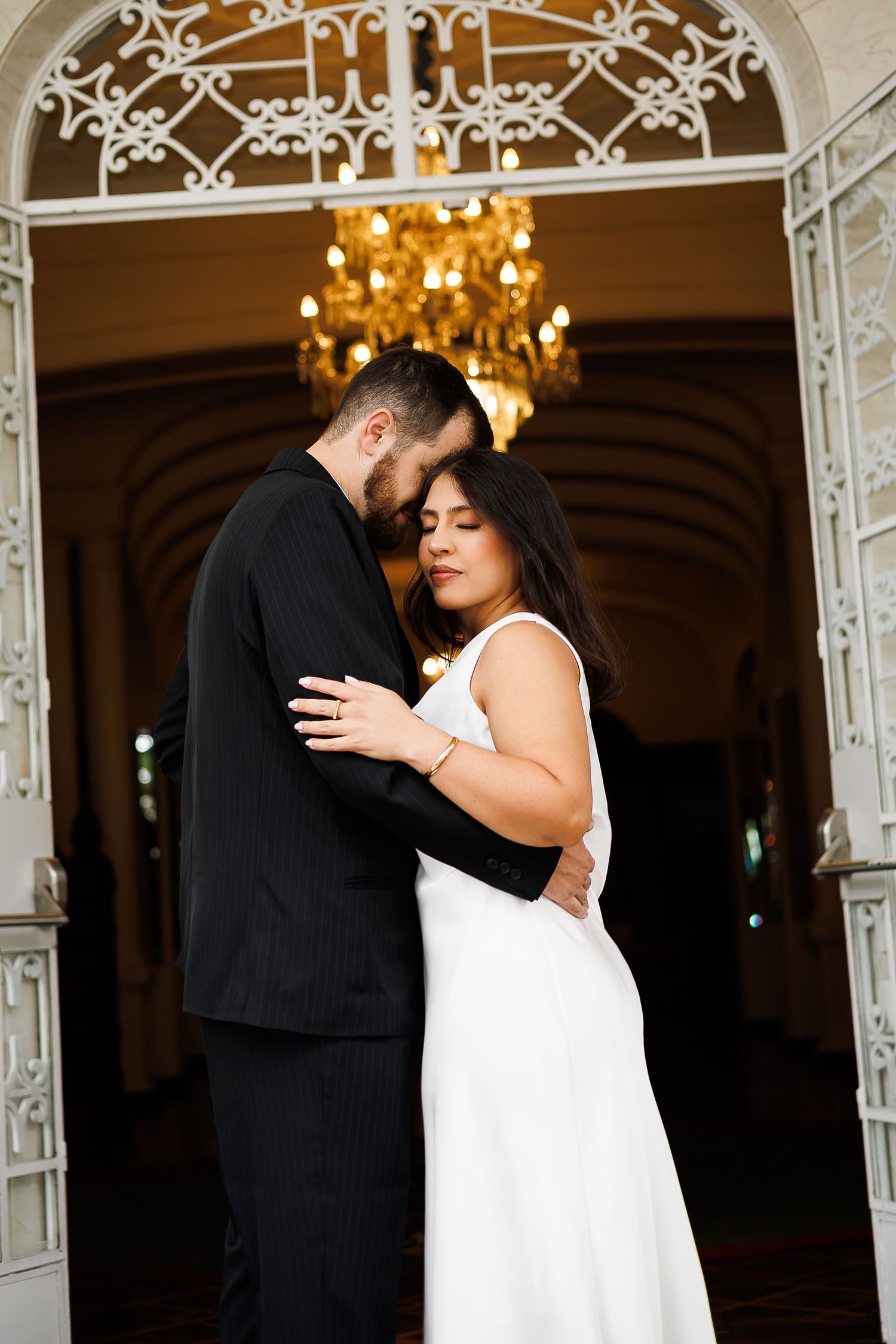 Bride and groom embrace tenderly in elegant venue with ornate white ironwork and glowing chandelier backdrop.