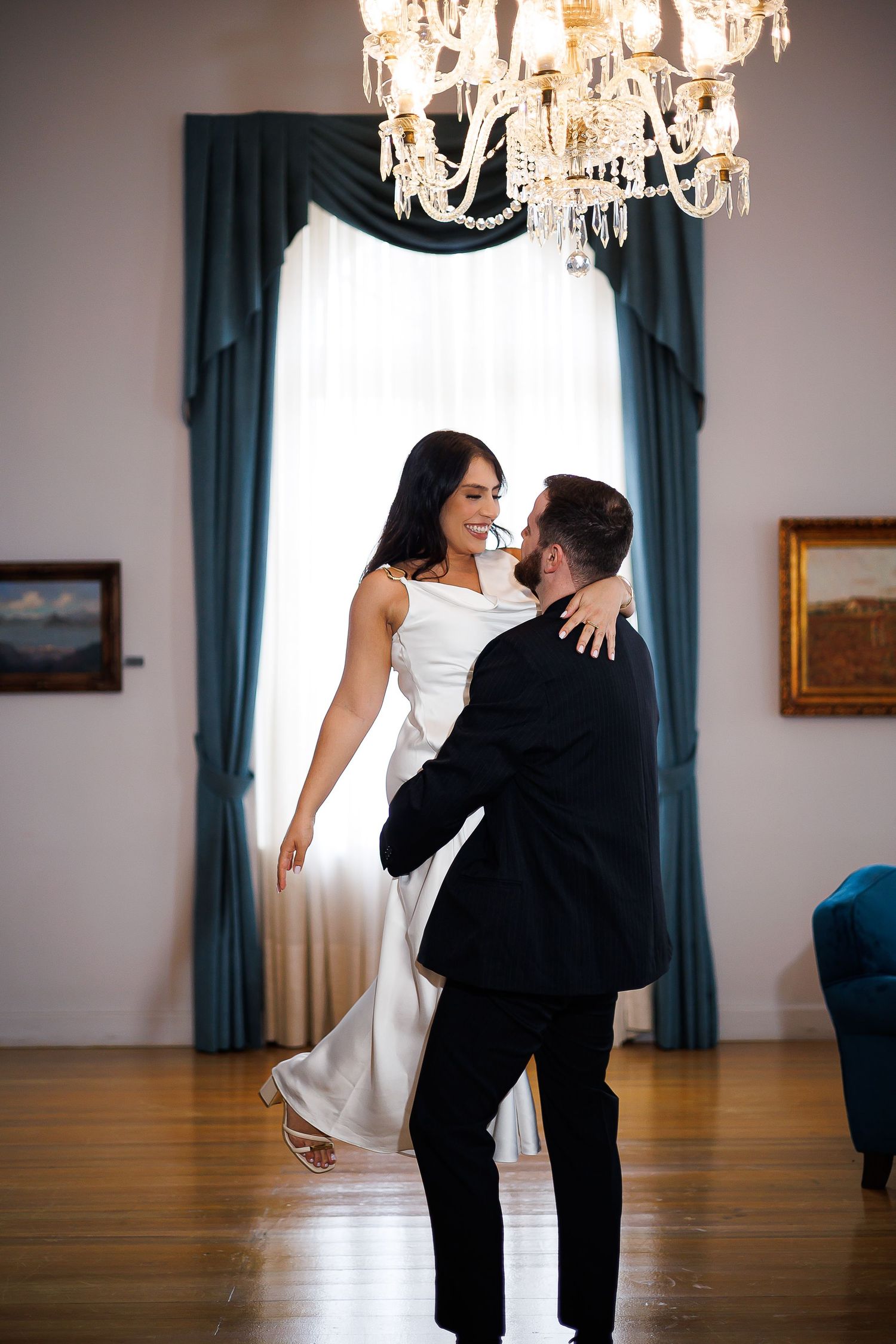 A bride and groom share a romantic first dance under a crystal chandelier in an elegant ballroom with teal drapes.