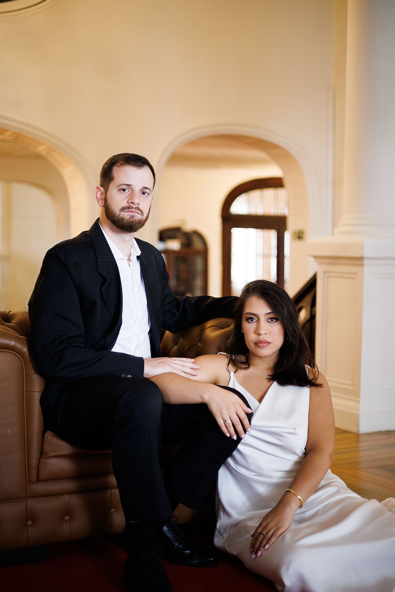 A man in a black suit and woman in a white dress pose elegantly in a grand interior with arched doorways.