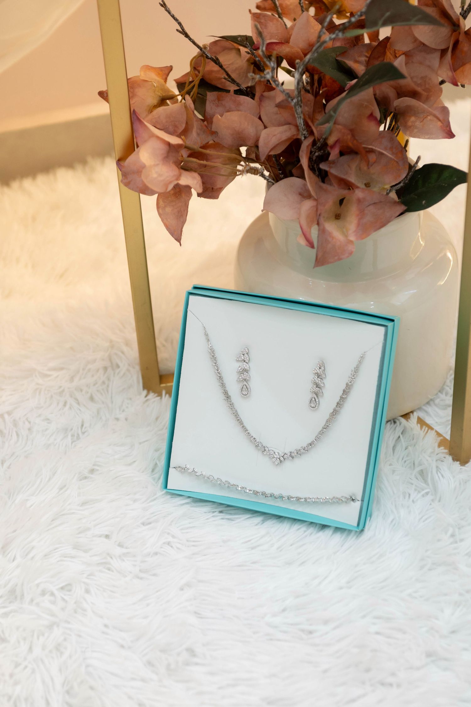 Silver crystal necklace and earring jewelry set displayed in teal gift box on white fur surface with floral decor.