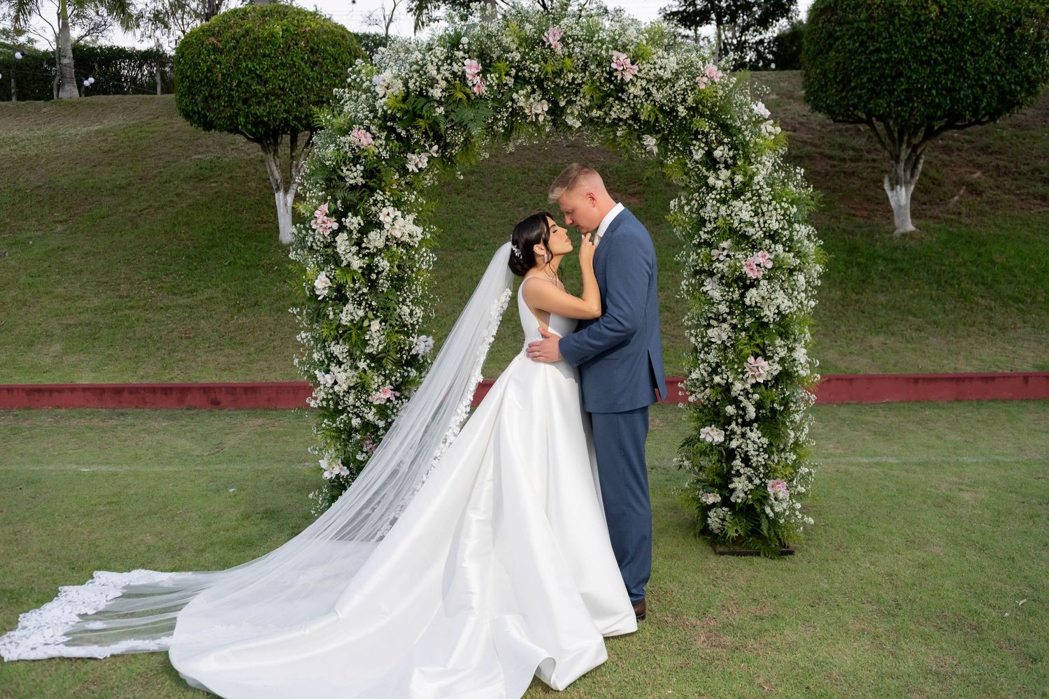 Bride and groom kissing under a floral arch on green lawn at outdoor wedding ceremony.
