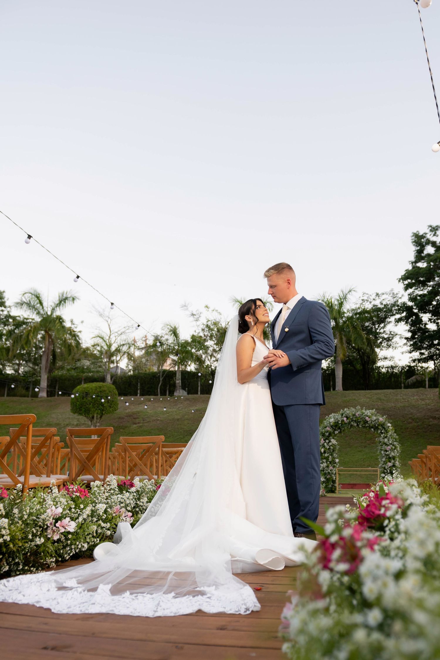 Bride and groom share a romantic moment at outdoor wedding ceremony with floral arrangements and string lights at dusk.