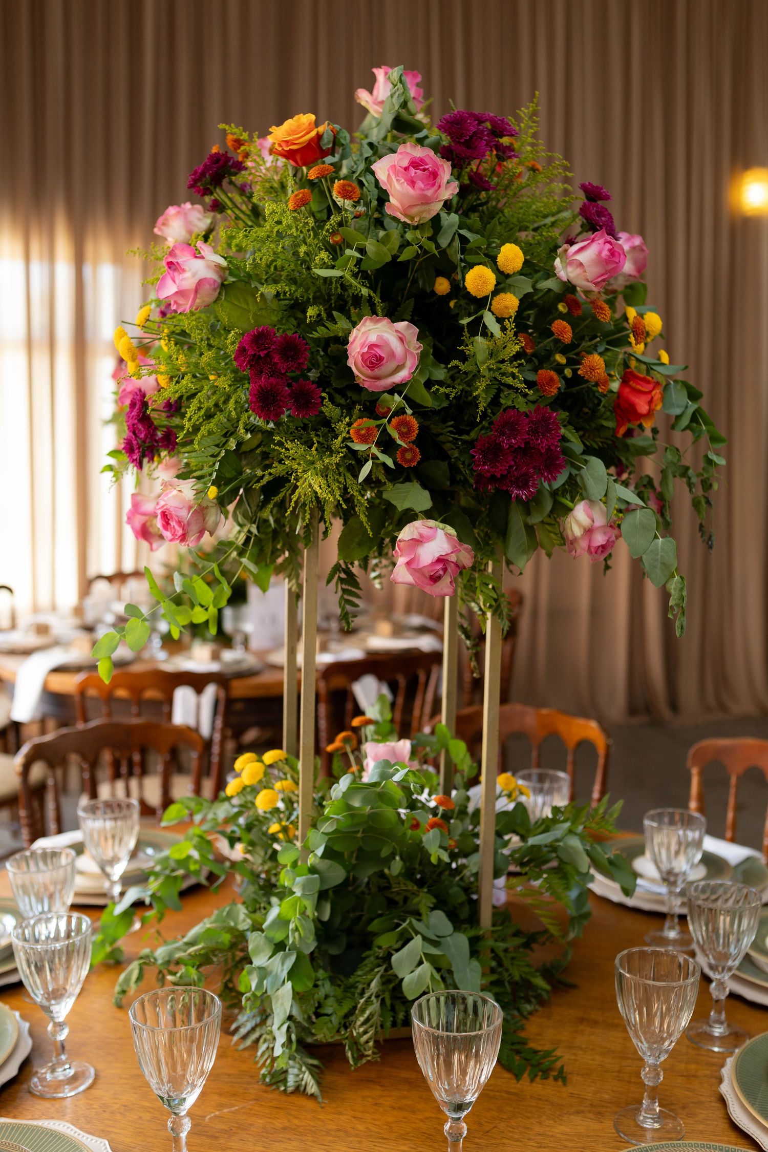 Tall floral centerpiece with pink, red, and orange roses on a wooden dining table set with crystal glasses.