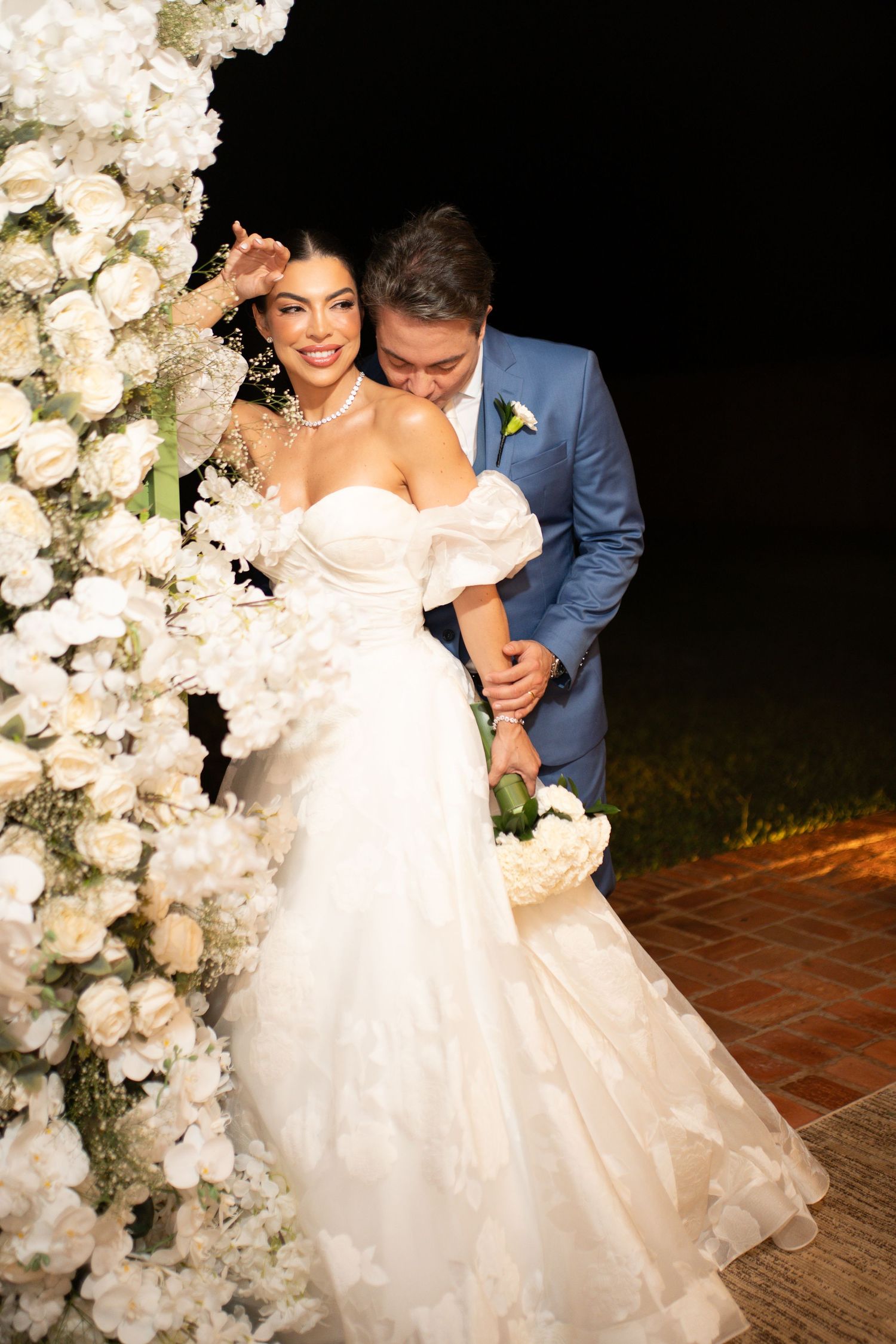 Bride in white off-shoulder gown holds bouquet as groom in blue suit kisses her cheek by white floral arch at night.