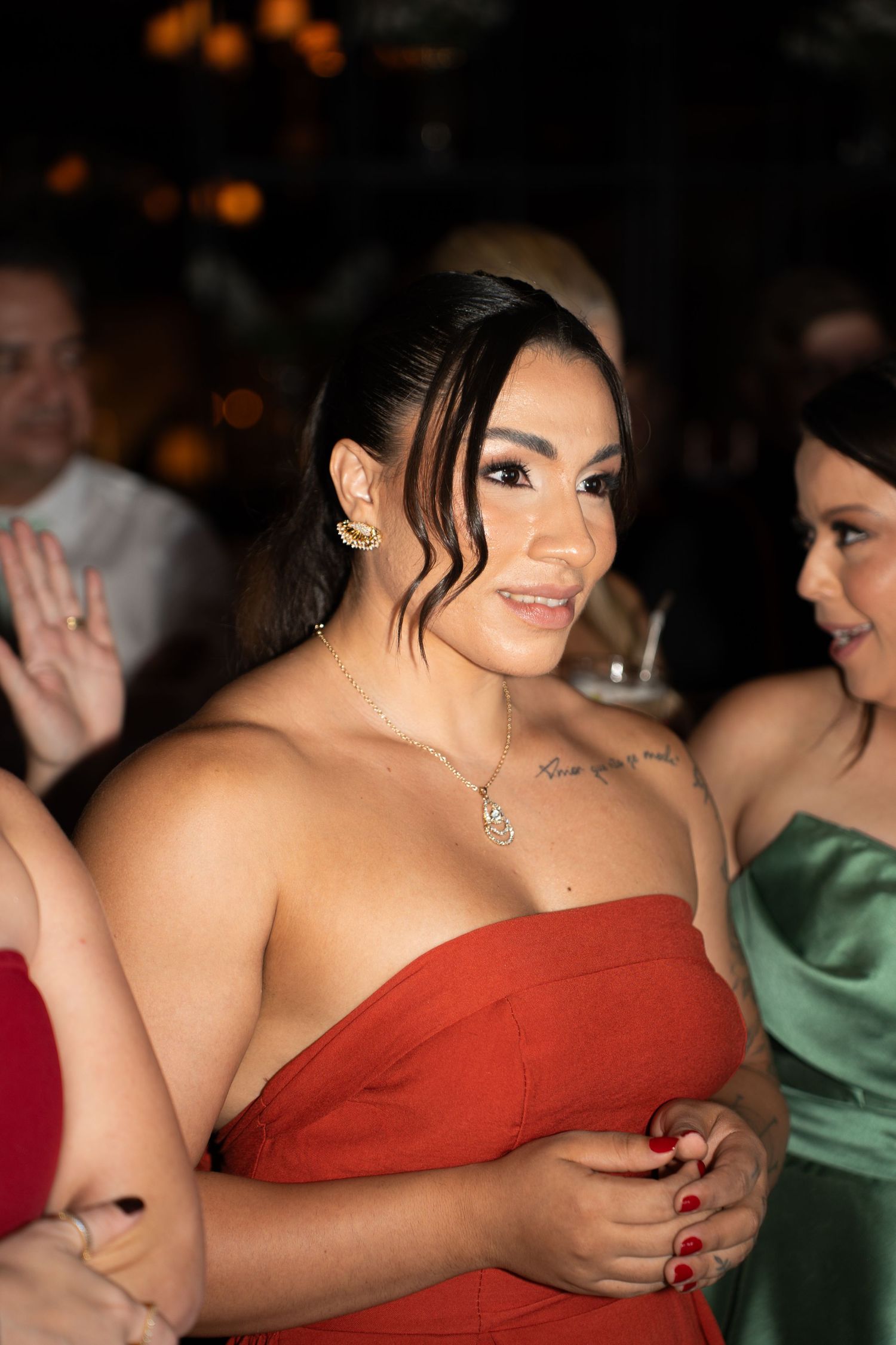 Woman in red off-shoulder dress with tattoo and gold jewelry smiling at formal event.