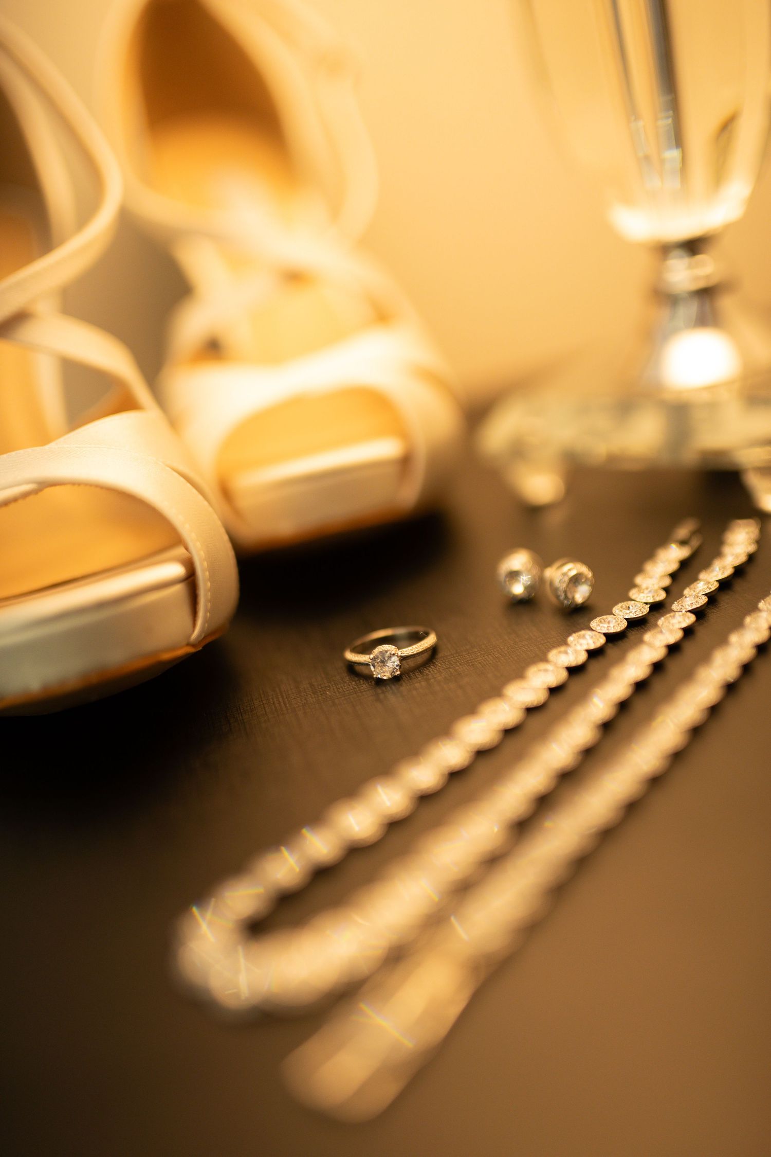 Bridal accessories flat lay featuring white peep-toe heels, diamond engagement ring, pearl necklace, and earrings on dark surface.