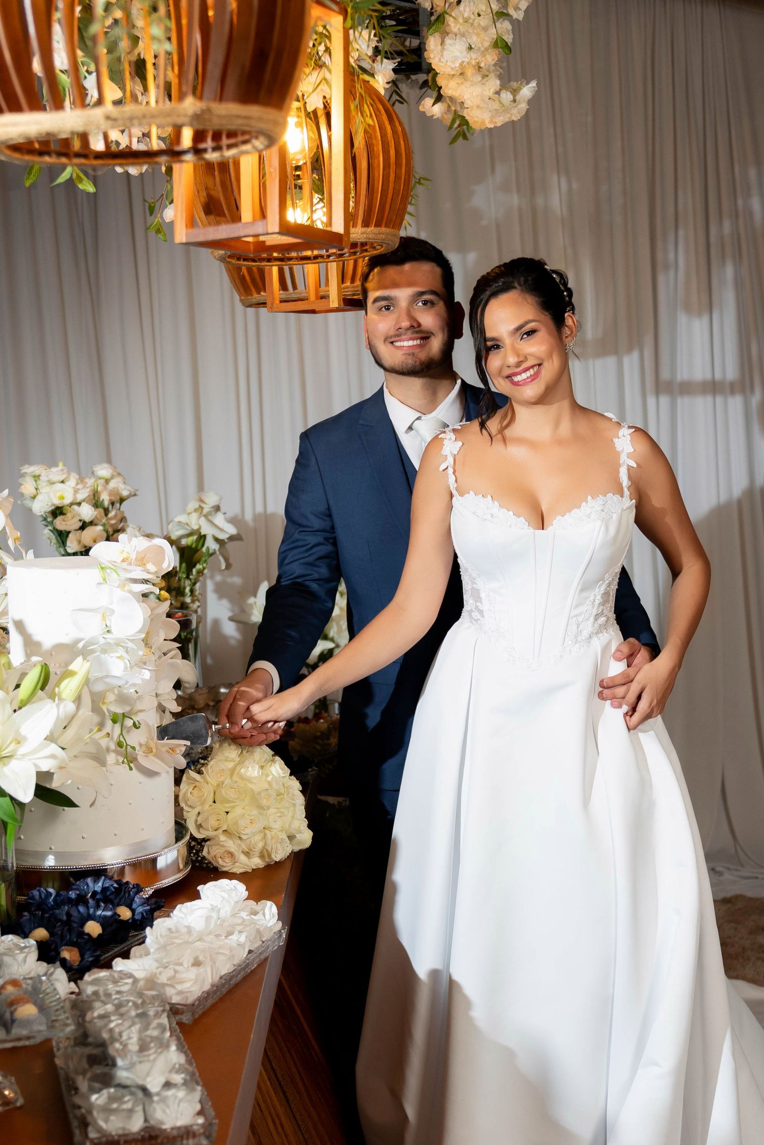 Smiling bride in white lace gown and groom in navy suit stand together at wedding cake table with floral decor.