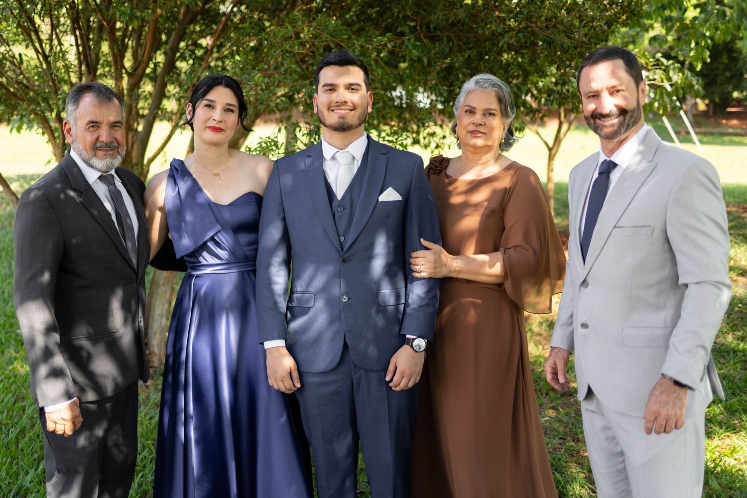 Group of five elegantly dressed people posing together outdoors at a formal event, smiling in a garden setting.
