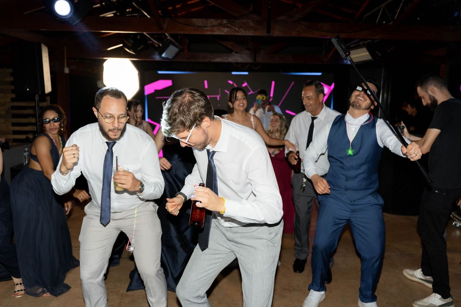Guests dancing energetically at a wedding reception in a rustic venue with colorful lights.