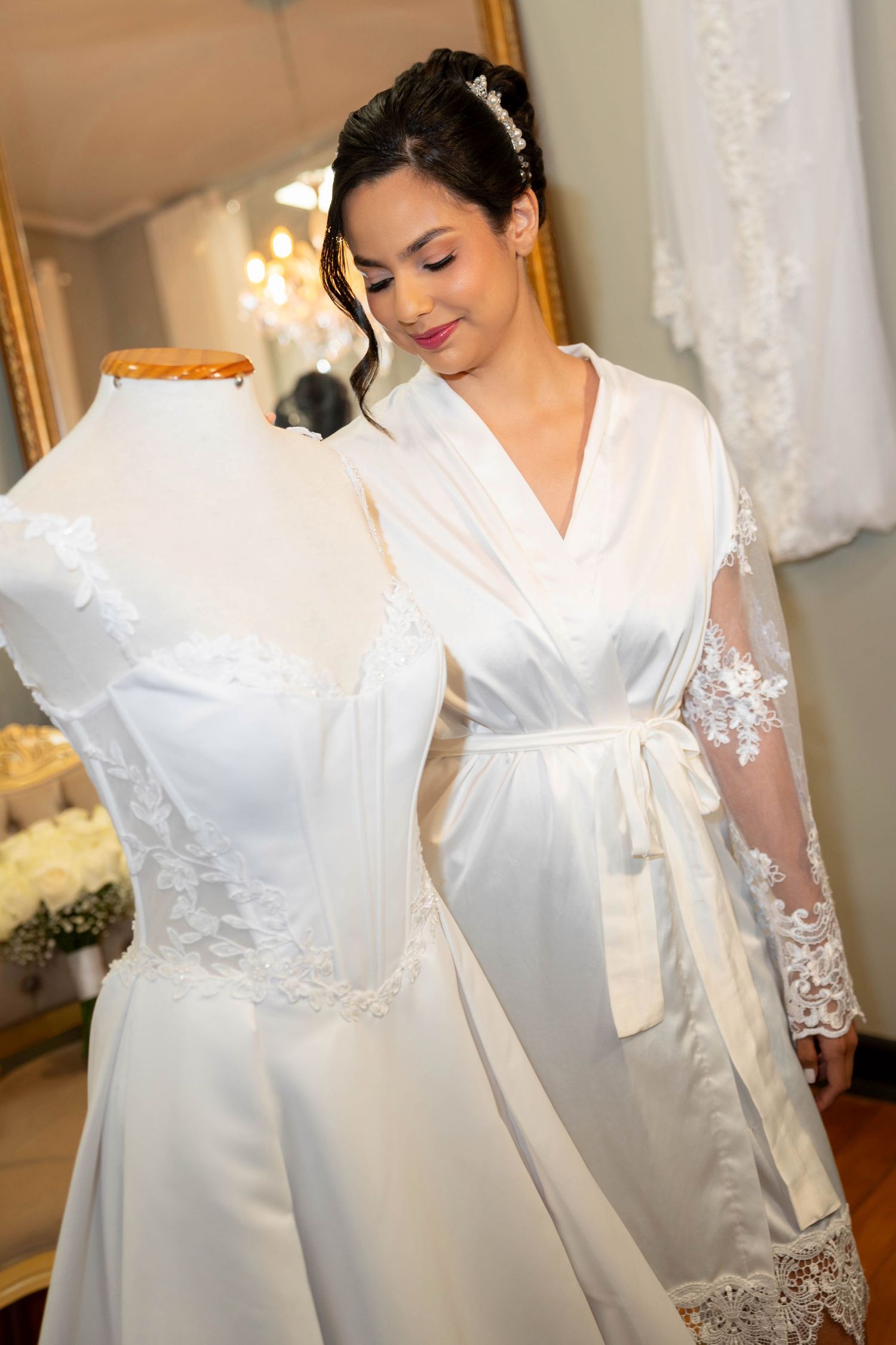 Bride in white satin robe admiring lace wedding dress on mannequin in bridal boutique.