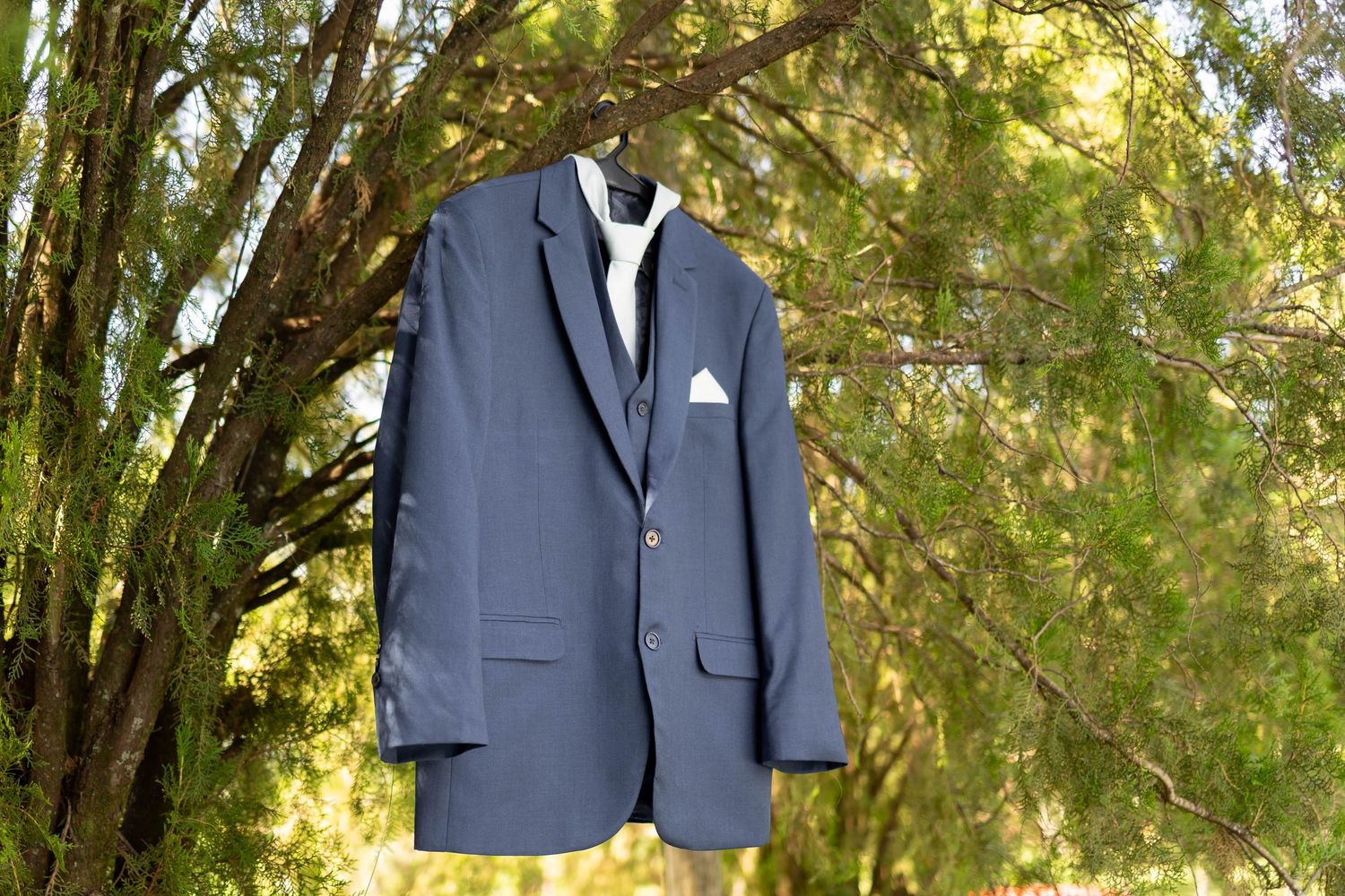 Navy blue suit jacket with white pocket square hanging on a tree branch outdoors.