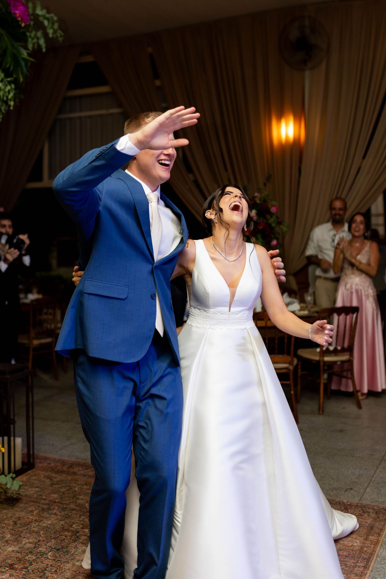 Joyful bride in white gown and groom in blue suit laughing during wedding reception entrance.