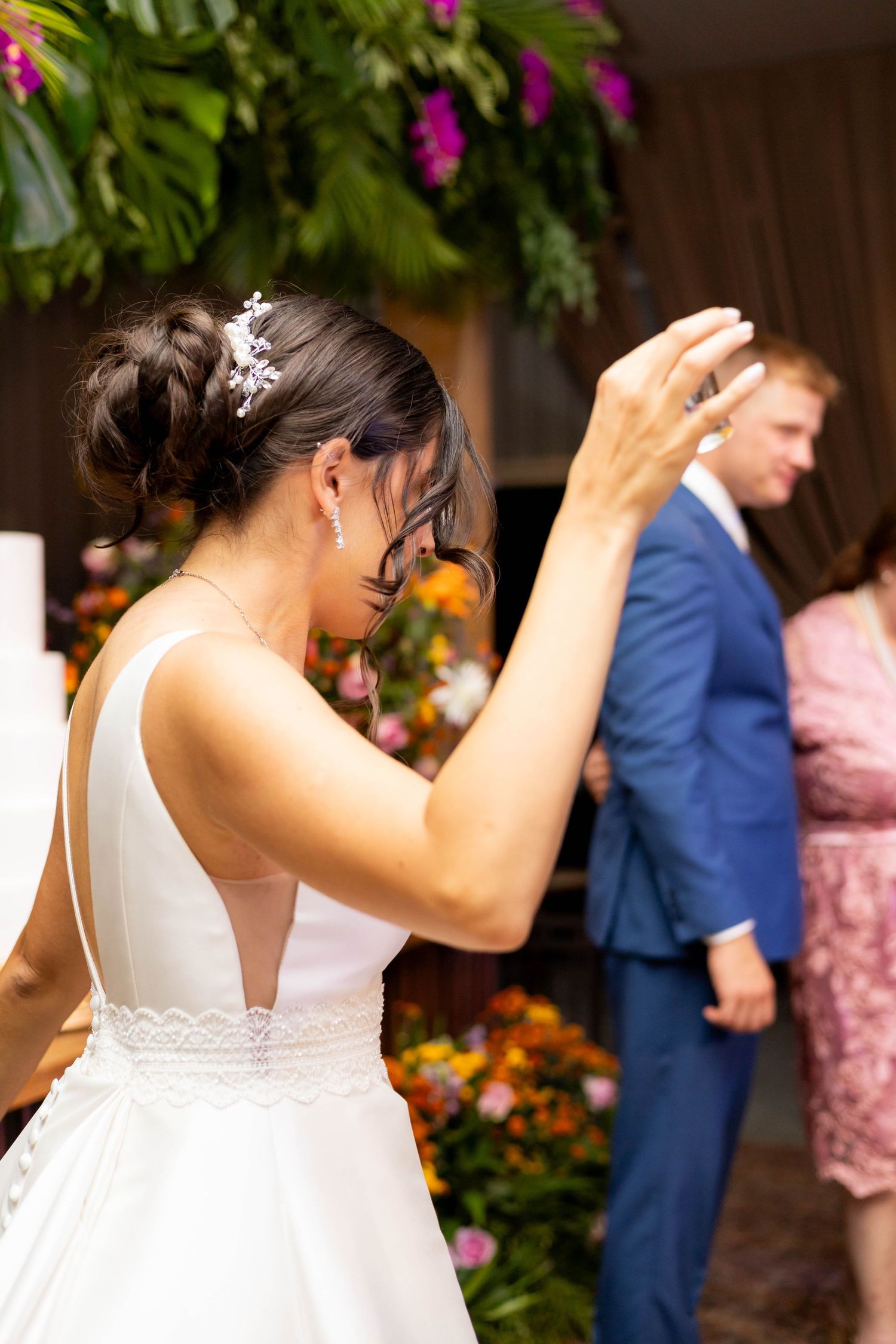 Bride in white gown with updo hairstyle raises hand during wedding ceremony with floral decorations.
