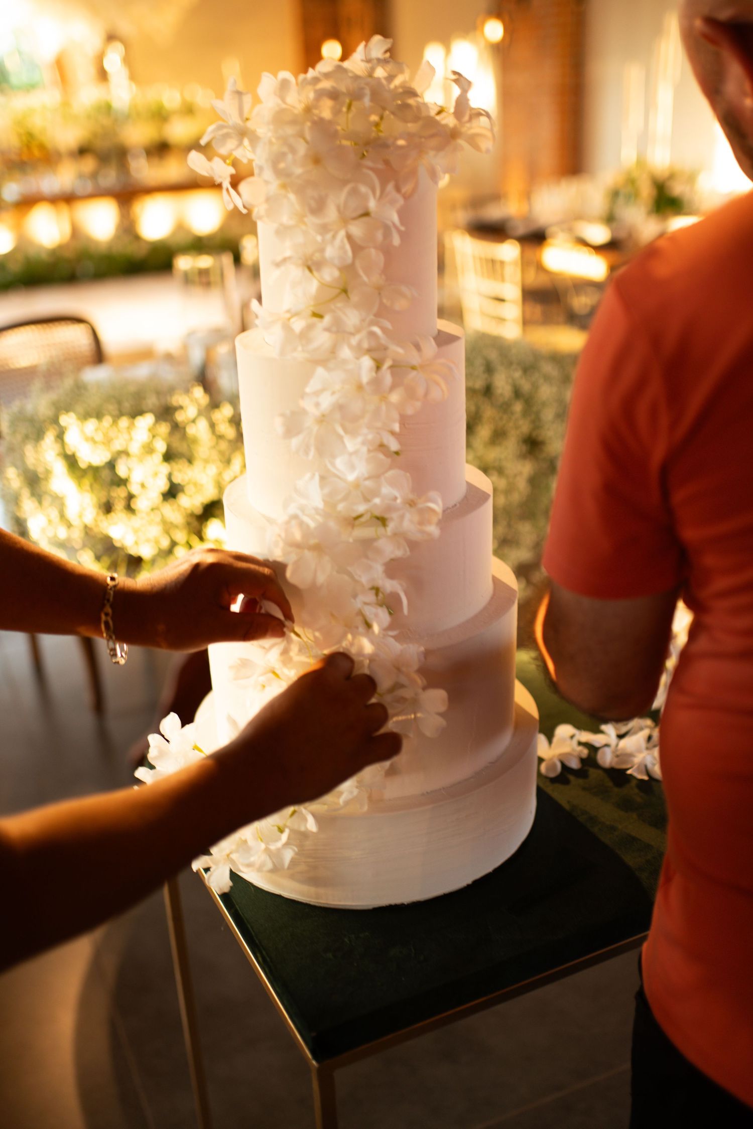 Elegant four-tier white wedding cake adorned with cascading white orchids, being decorated by hands at a reception venue.