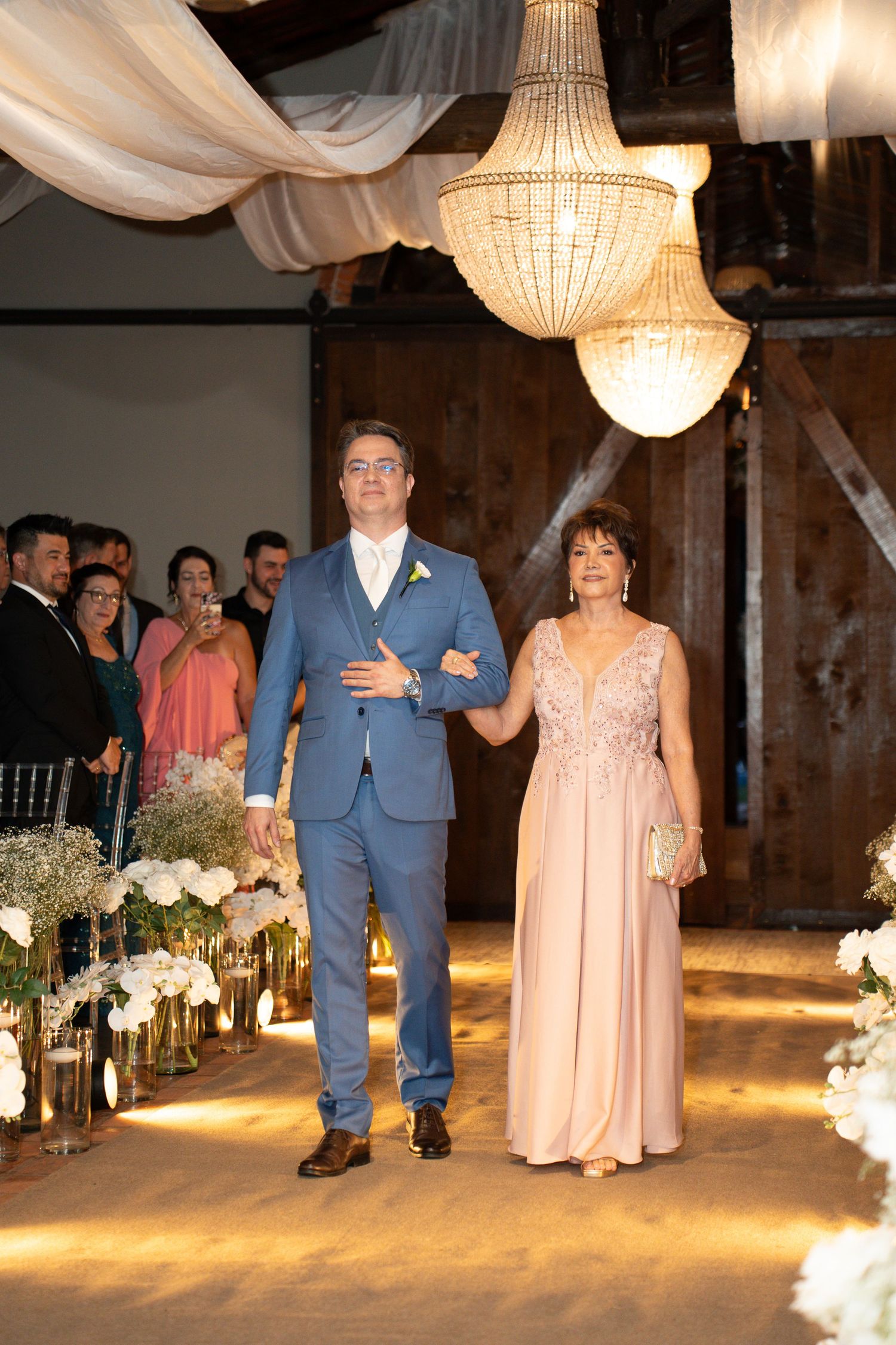 A couple walks down a wedding aisle in an elegant barn venue with chandeliers, the man in blue suit and woman in pink gown.
