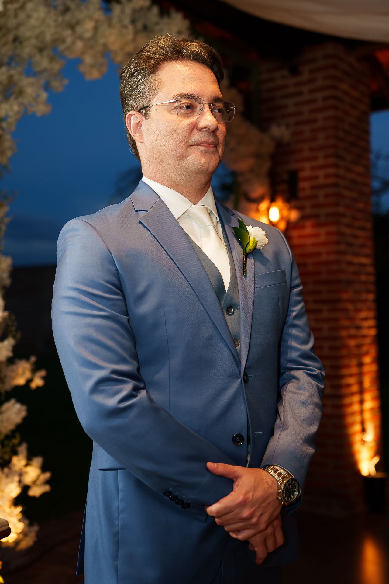 A groom in a blue suit with boutonniere stands confidently at the altar during an elegant evening wedding ceremony.