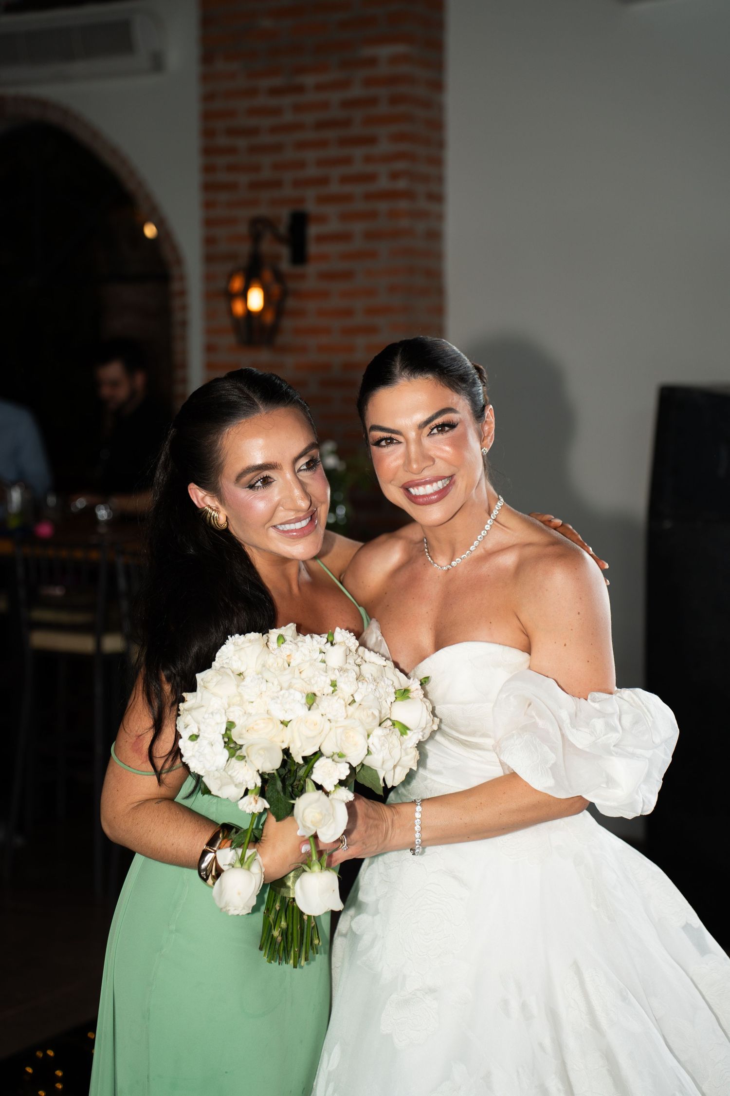 Bride in white off-shoulder gown holds white rose bouquet with bridesmaid in green dress at indoor wedding reception.