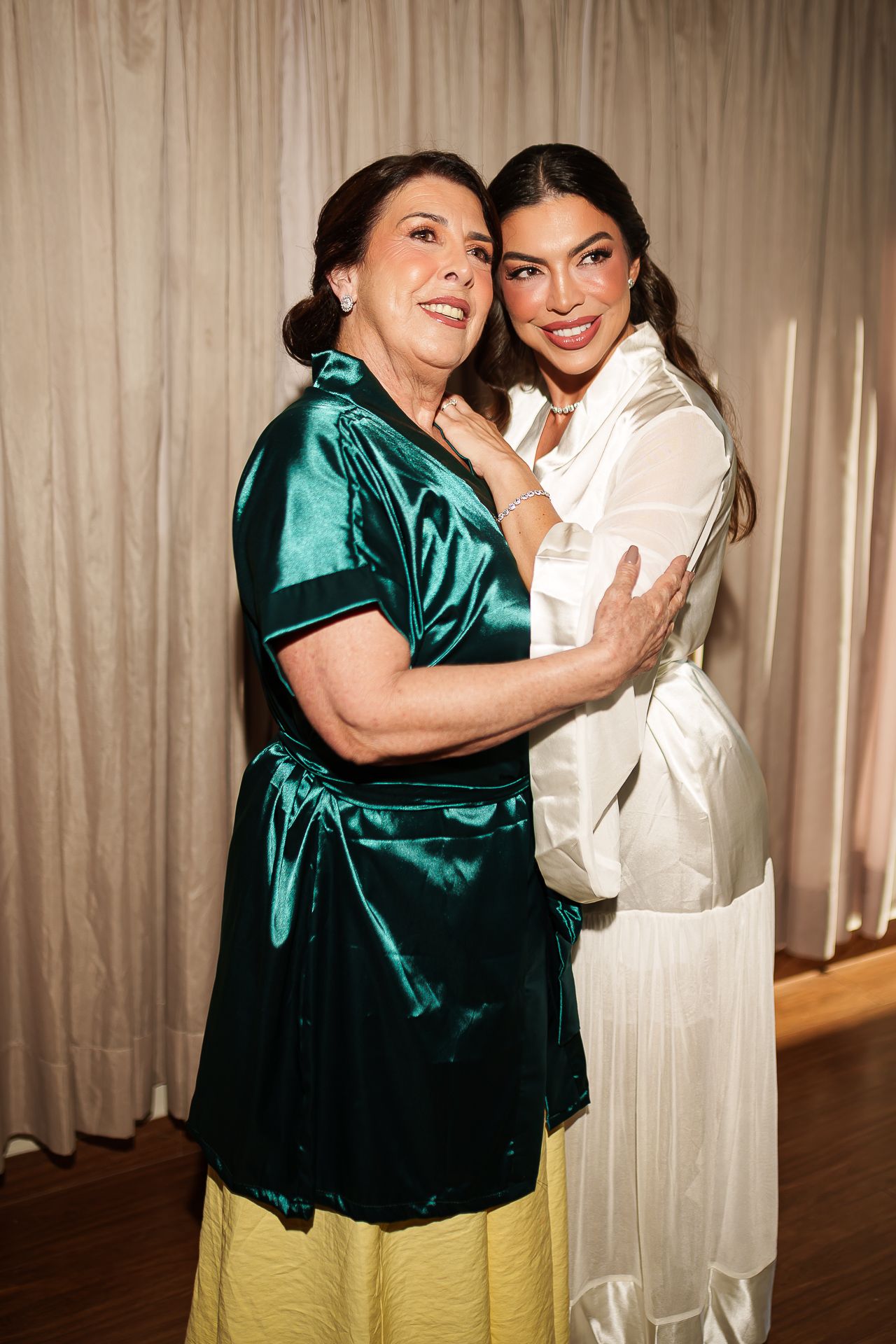 Two women in elegant satin outfits embracing and smiling warmly in a softly lit indoor setting.