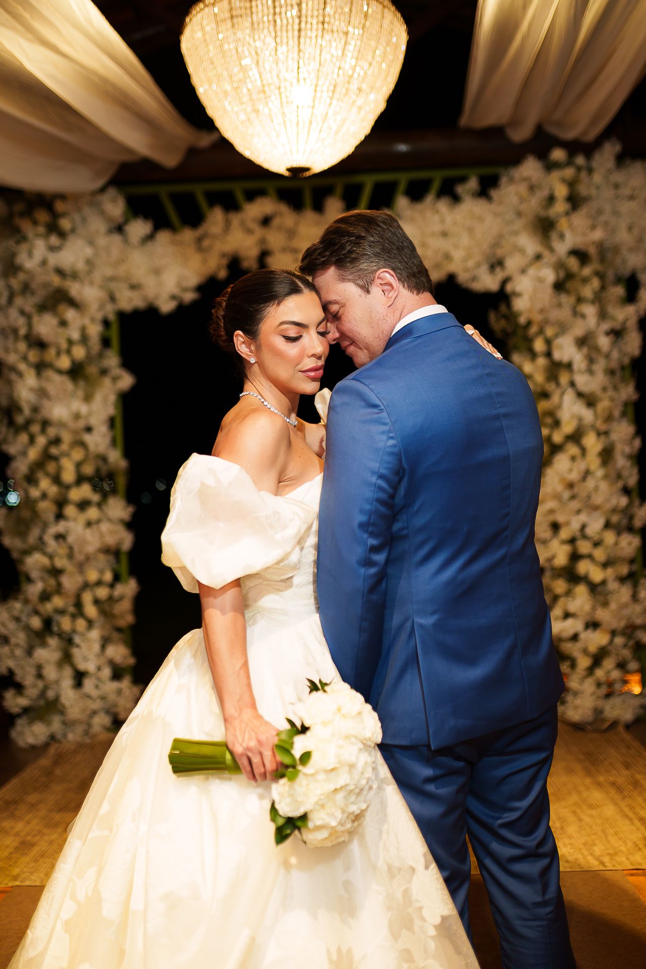 Bride in off-shoulder white gown holds bouquet as groom in blue suit leans in tenderly before floral backdrop.