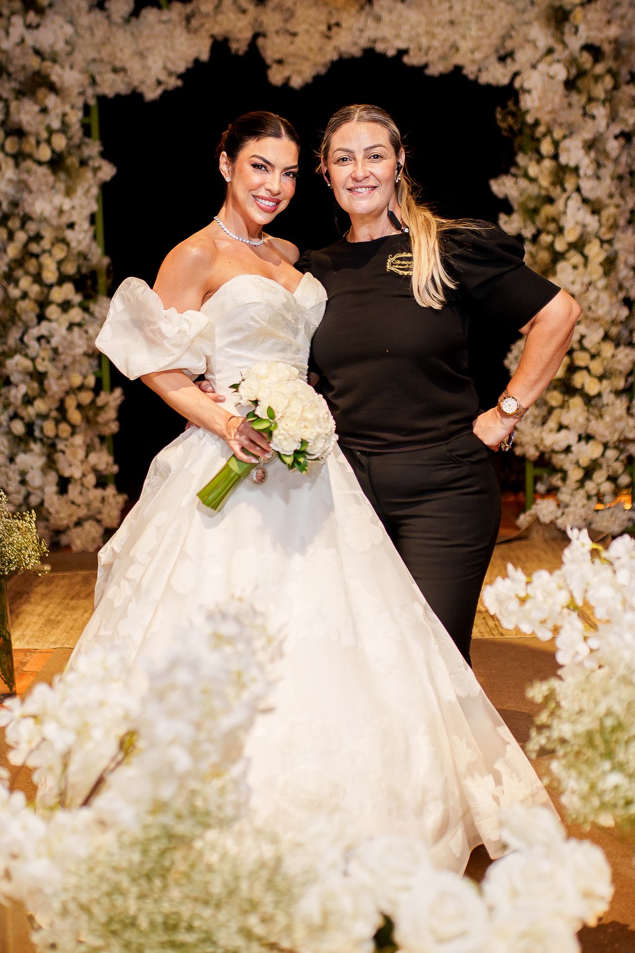 Bride in white ballgown holding bouquet poses with woman in black dress amid white floral wedding decor.