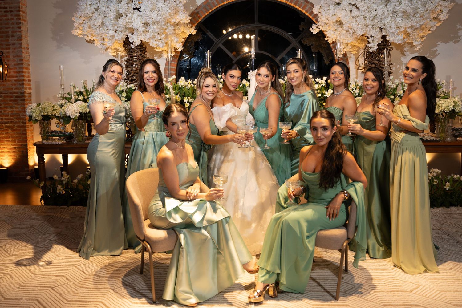 Bride poses with bridesmaids in sage green satin dresses, holding champagne glasses at elegant floral wedding venue.
