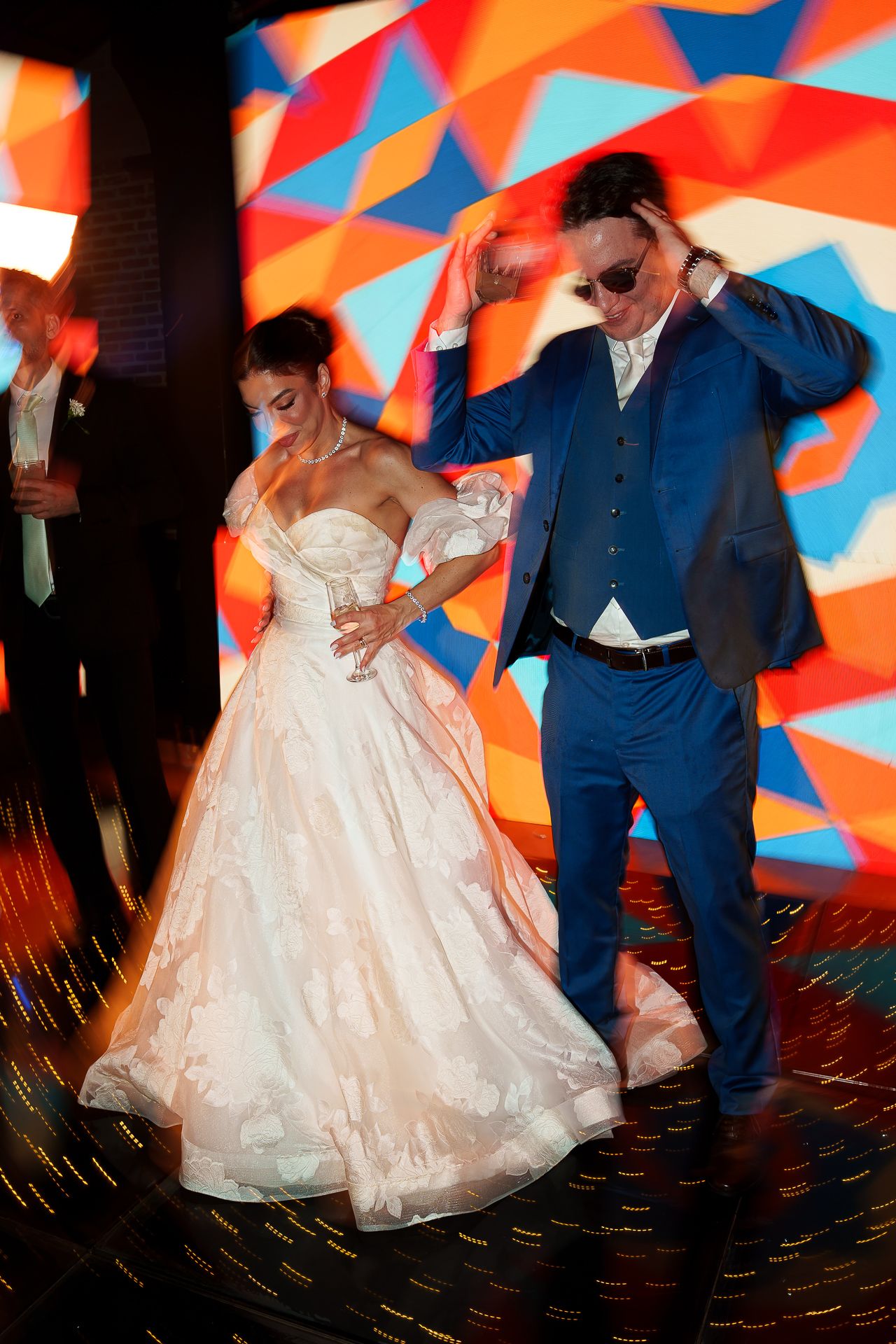 Bride in white gown and groom in blue suit dancing energetically at wedding reception with colorful geometric backdrop.