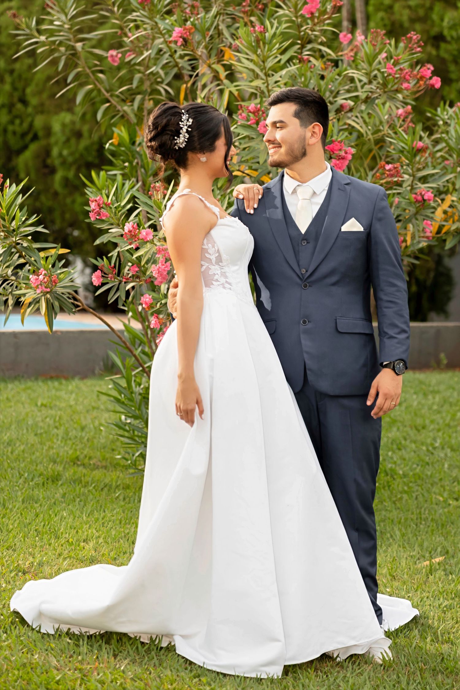 Bride in white gown and groom in navy suit smiling together outdoors with pink flowers.