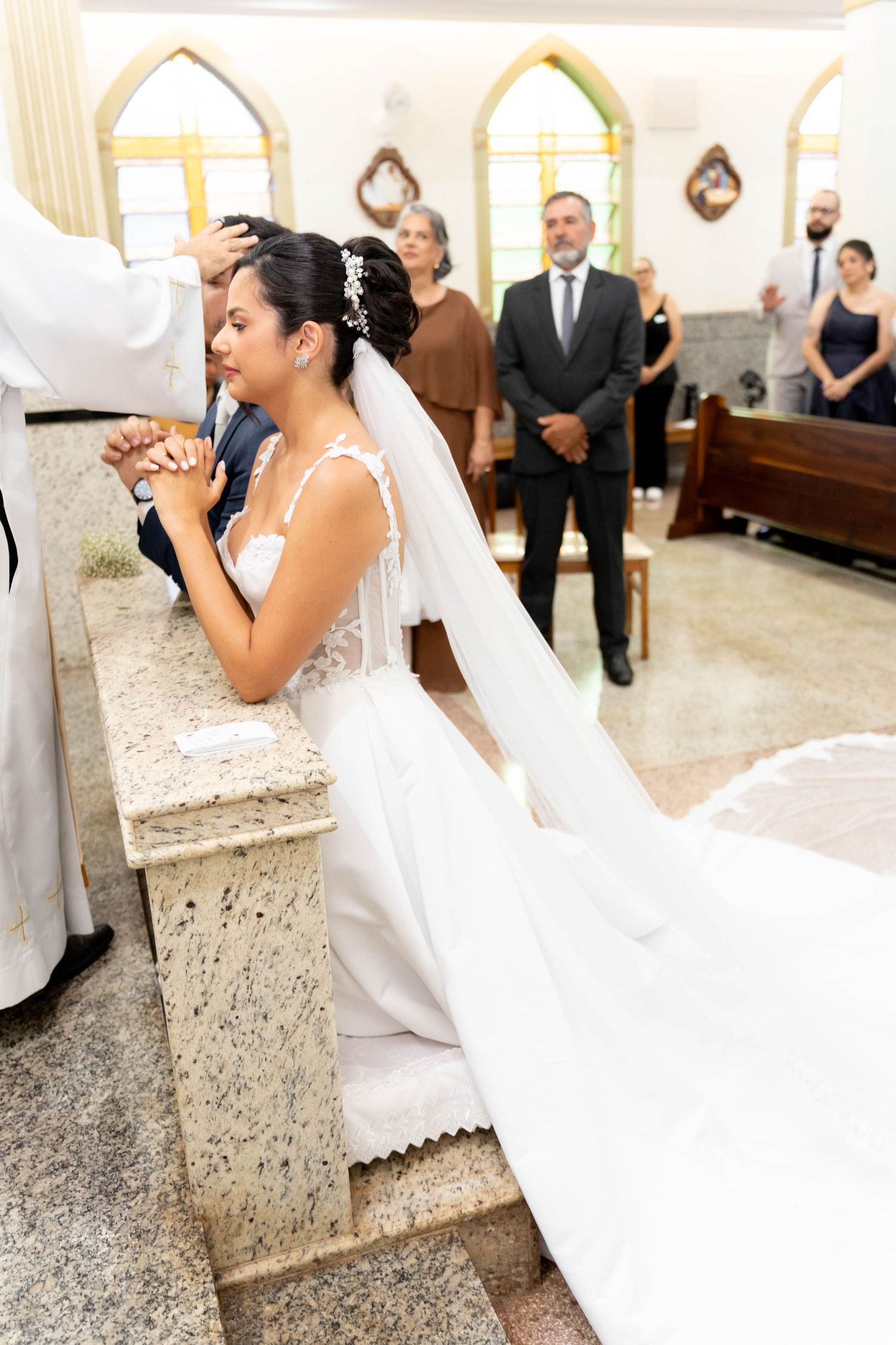 Bride kneeling at church altar in white gown and veil during wedding ceremony with guests standing behind.