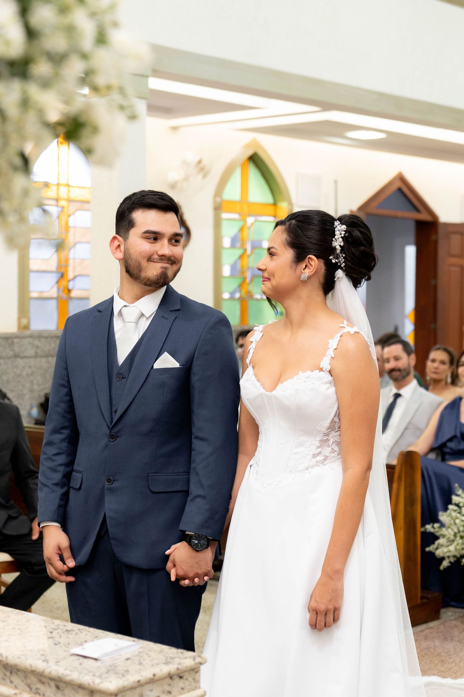 Bride and groom holding hands at church altar, she in white lace gown, he in navy suit, during wedding ceremony.