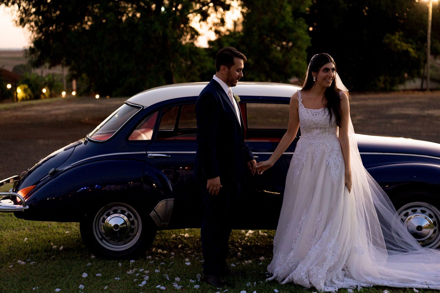 Bride and groom holding hands beside a classic vintage navy blue car at dusk during outdoor wedding photo session.