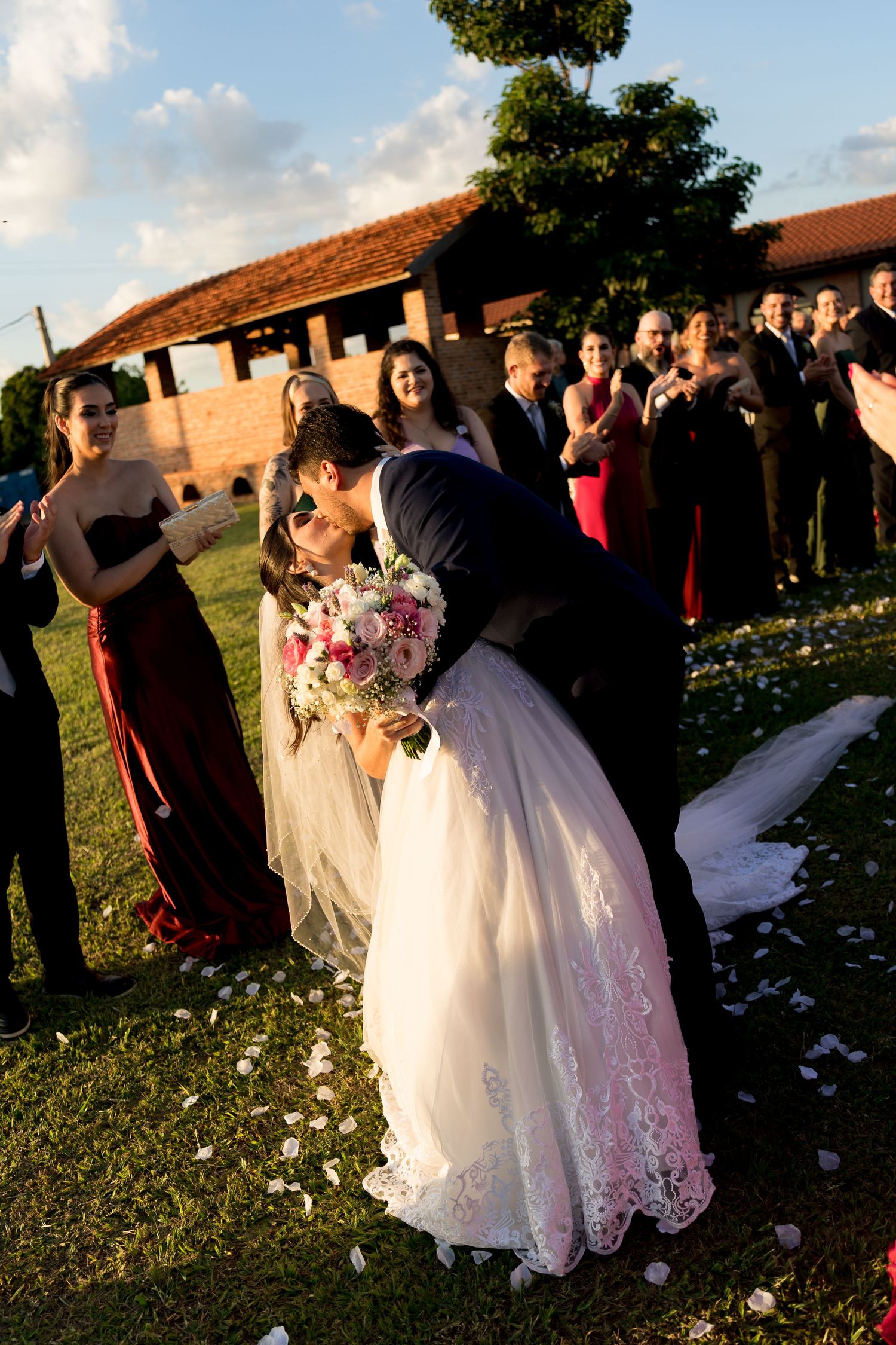 A bride and groom share a romantic kiss outdoors at their wedding reception while guests celebrate around them.