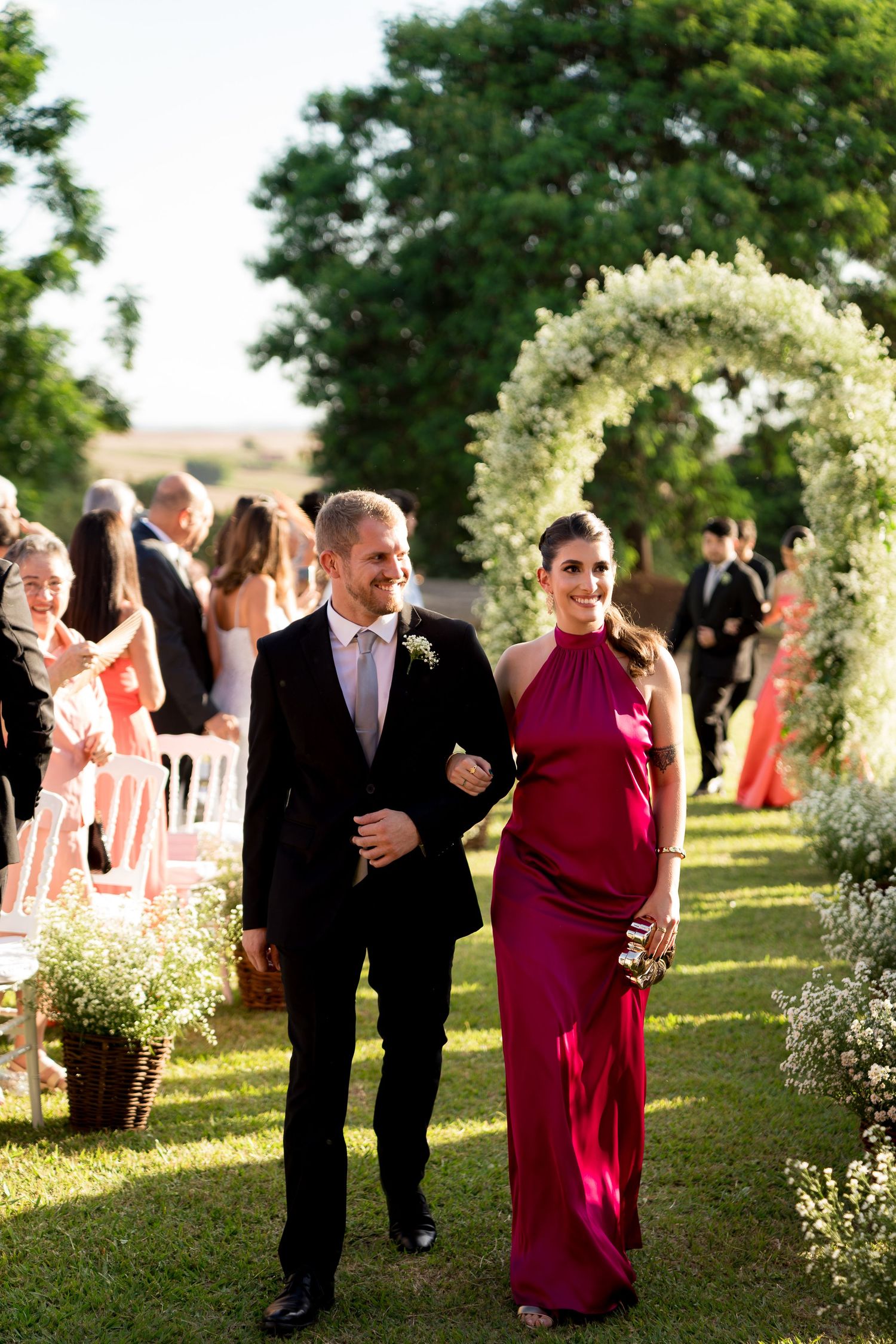 Couple walks down outdoor wedding aisle, man in black suit, woman in red gown, floral arch backdrop.