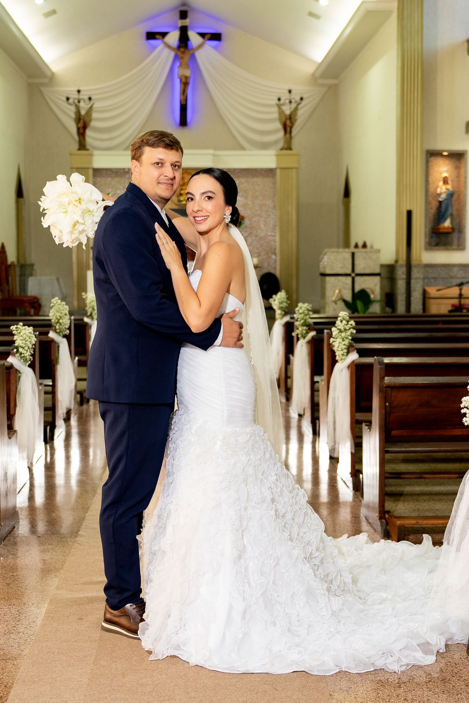 Bride and groom embracing in a decorated Catholic church aisle, bride in white mermaid gown with veil, groom in navy suit.