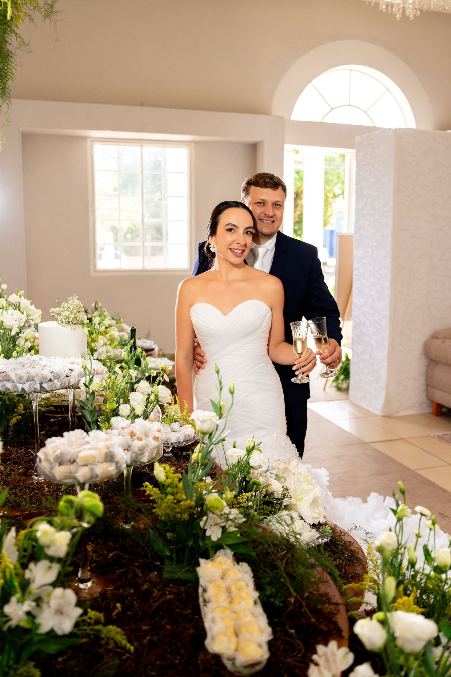 Bride and groom smiling together at wedding reception, surrounded by white floral arrangements and champagne glasses.