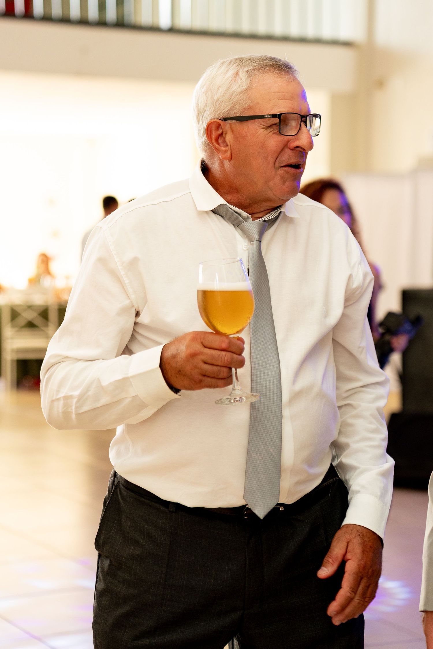 Elderly man in white shirt and gray tie holding a glass of beer at a formal event.