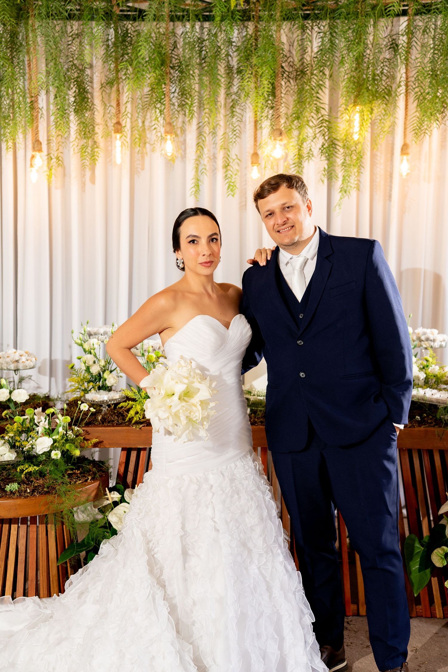 Bride in white ruffled gown holding bouquet stands with groom in navy suit at elegant wedding reception.